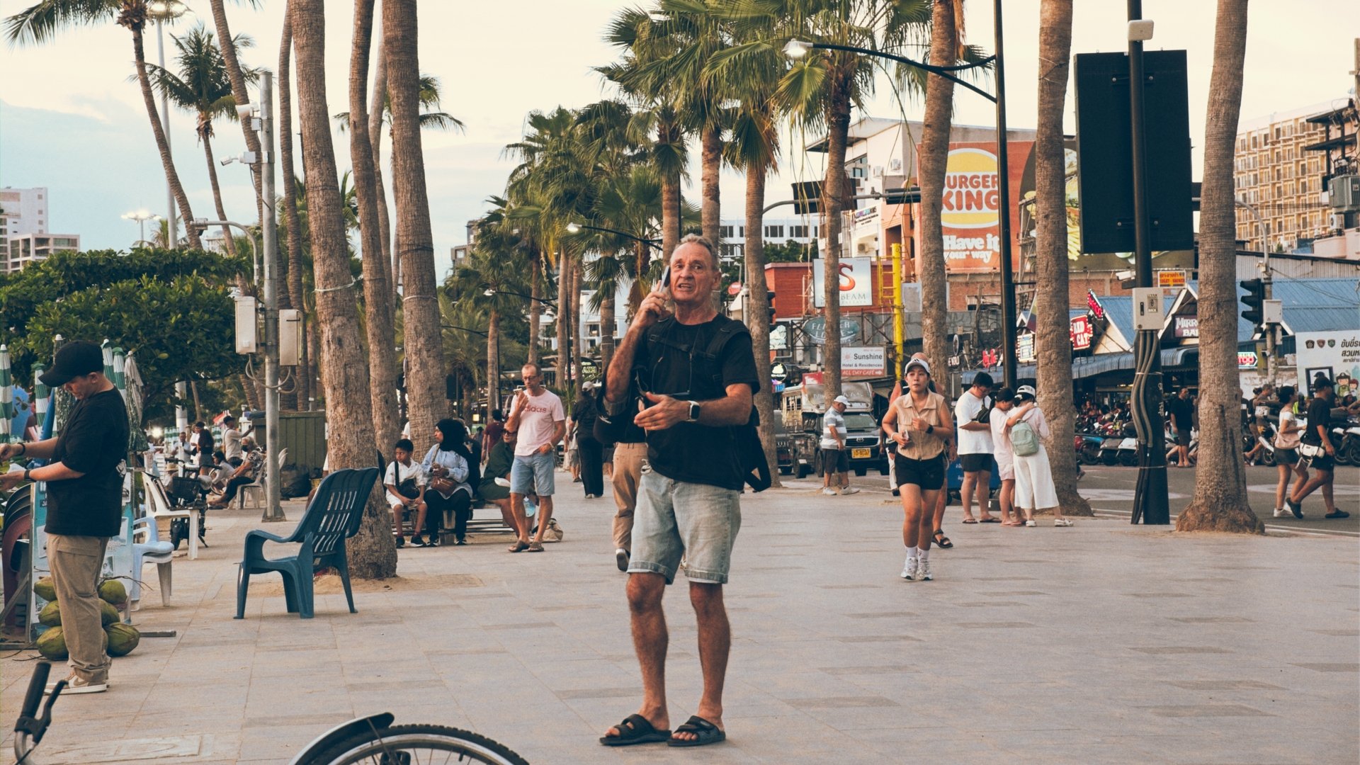 People walking and sitting in an urban outdoor area lined with palm trees, including a man in shorts and flip-flops talking on a cell phone in the foreground, with various shops, signs, and seated individuals in the background.