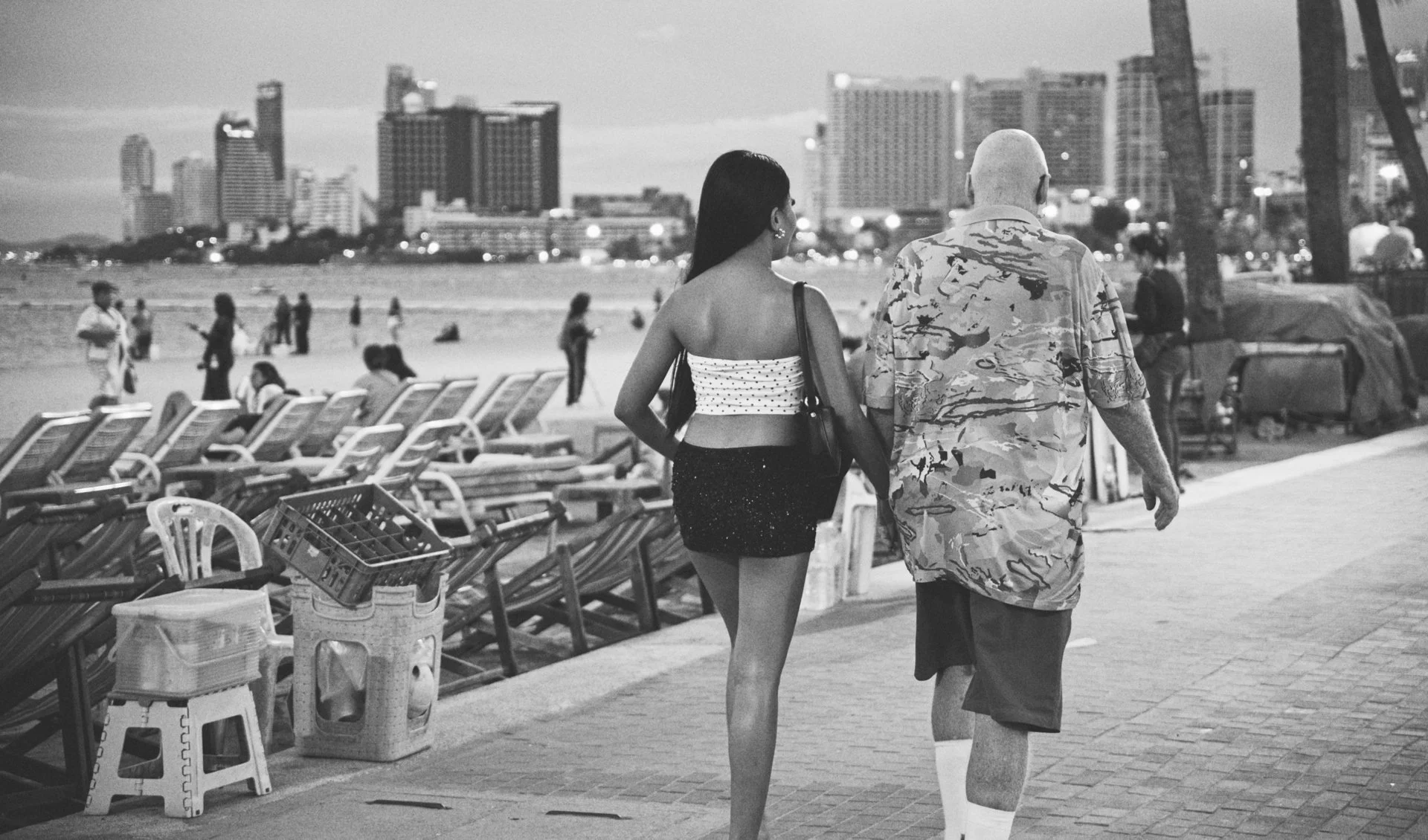 A black-and-white photograph of a man and woman walking along a beach promenade. The woman is wearing a strapless top and a short skirt, and the man is wearing a camouflage shirt and shorts. In the background, there are beach chairs, a city skyline w