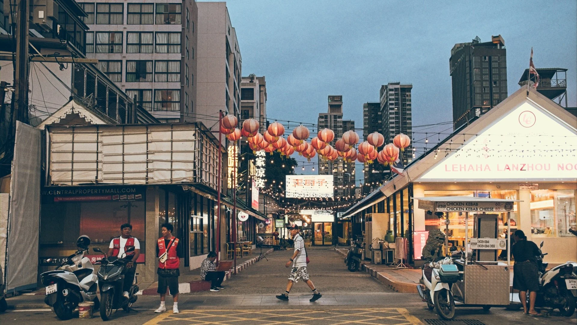 A busy street scene in an Asian city during evening with hanging red lanterns and modern high-rise buildings in the background. There are shopfronts with signs, scooters parked, and people walking.