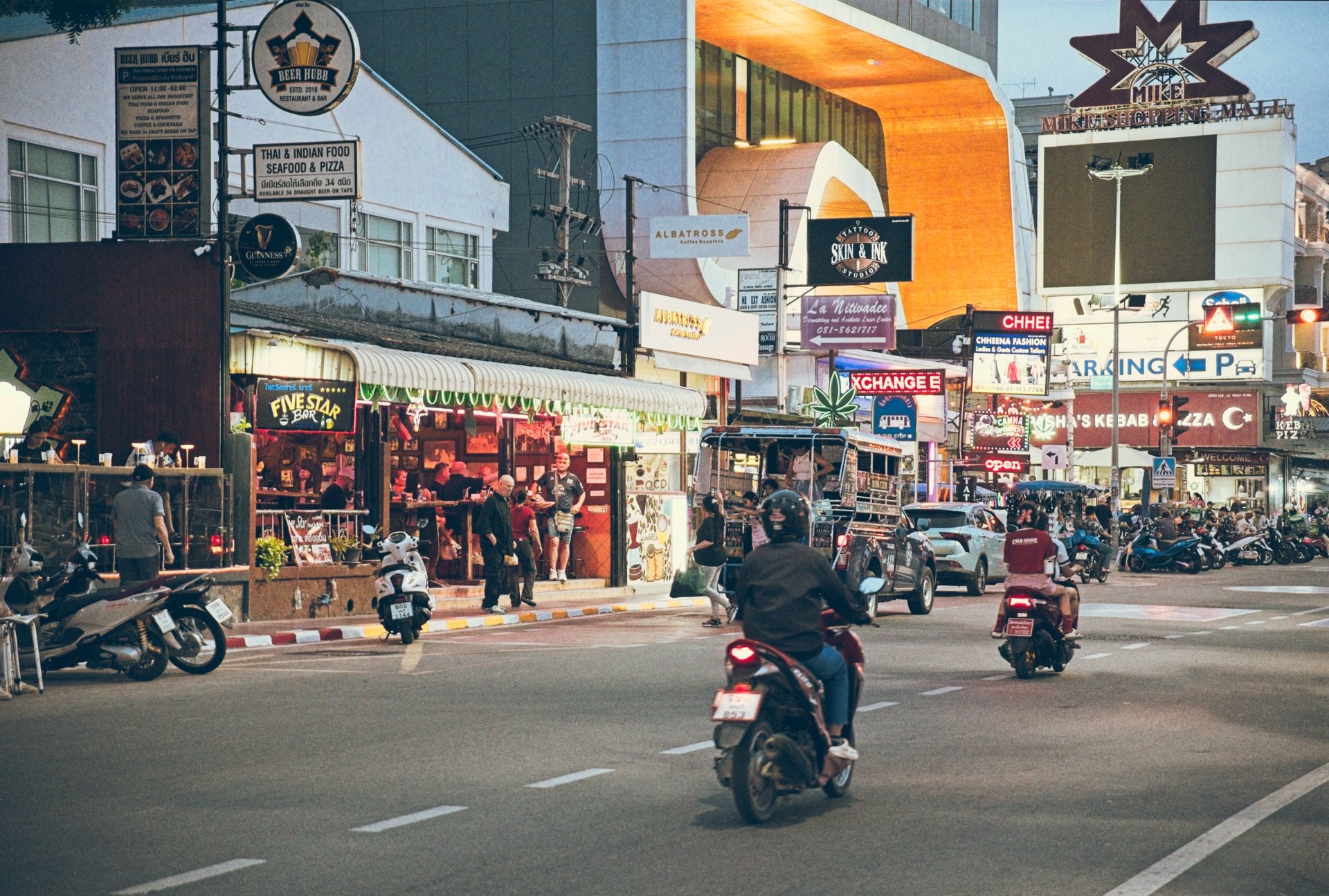 City street scene with people riding motorcycles, parked scooters, shops, and illuminated signs. Buildings line the street with restaurant and business signs visible.