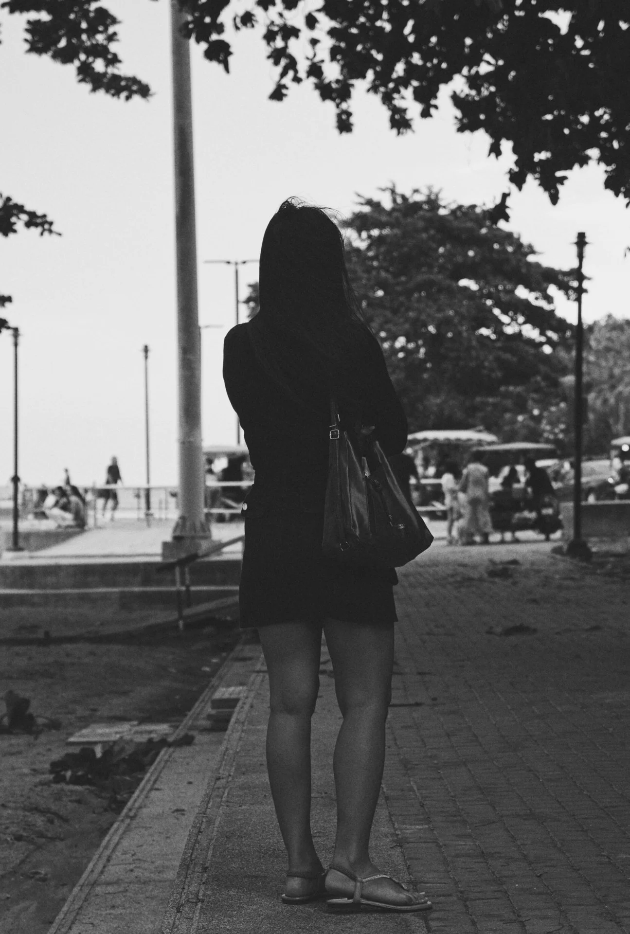 A black and white photo of a woman standing outdoors, with trees and people in the background. She is facing away from the camera, wearing a dress, sandals, and carrying a handbag.