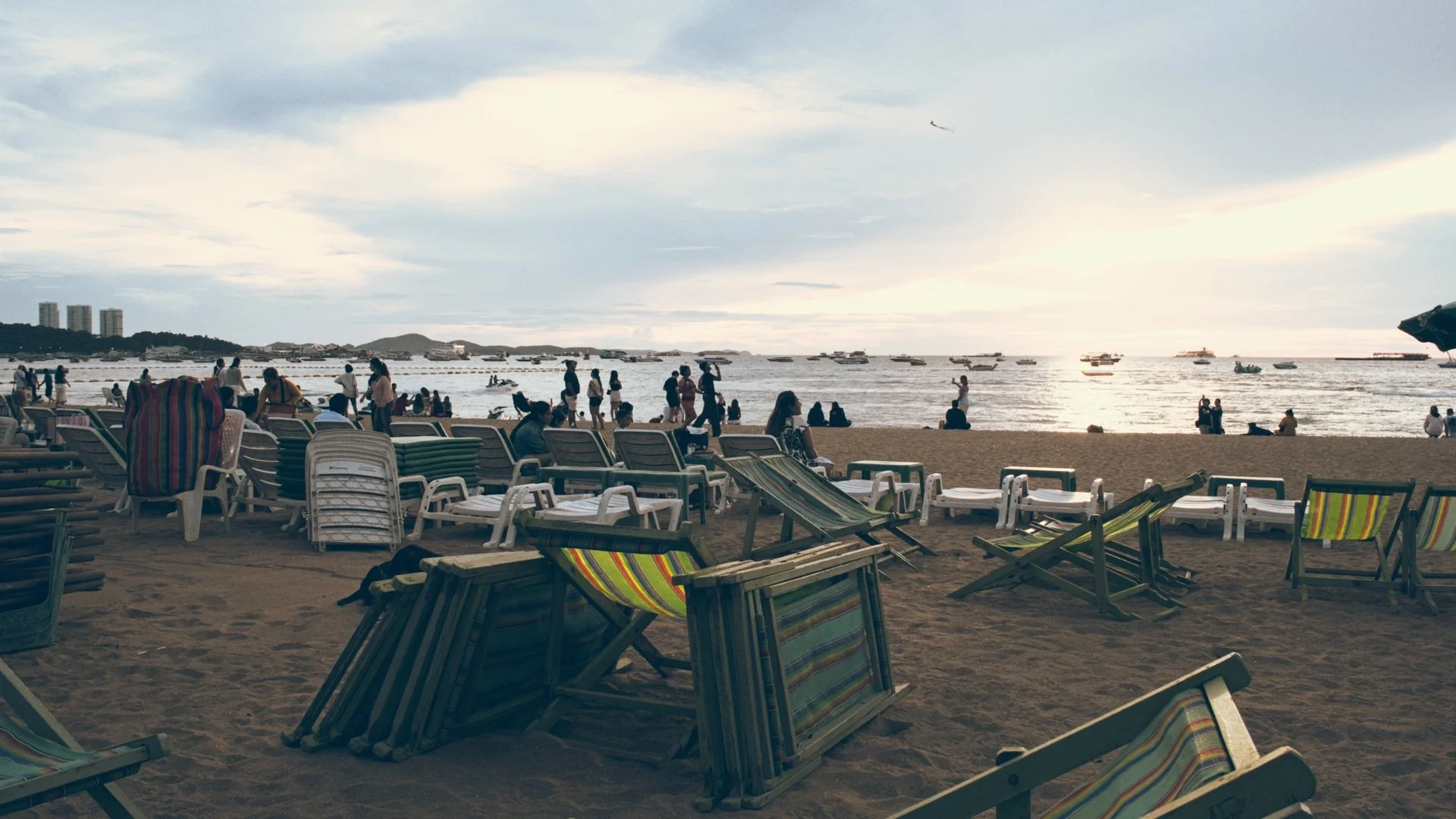 Beach scene in the late afternoon with numerous lounge chairs, some occupied by people, facing the ocean. Many people are walking or sitting on the sand, and boats are anchored offshore. The sky is partly cloudy with some sunlight breaking through.