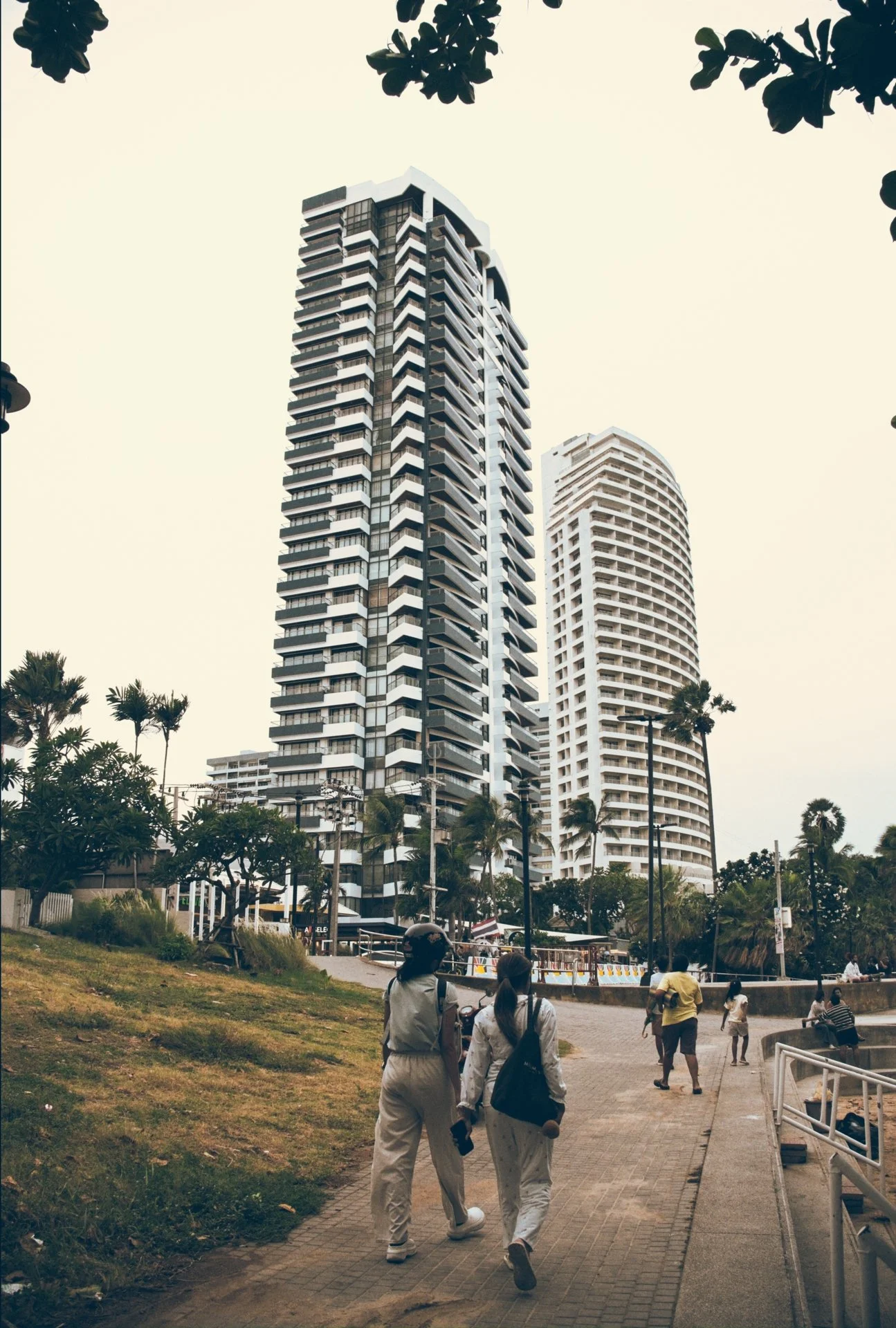 People walking along a paved pathway with green grass, trees, and tall modern buildings in the background, under an overcast sky.