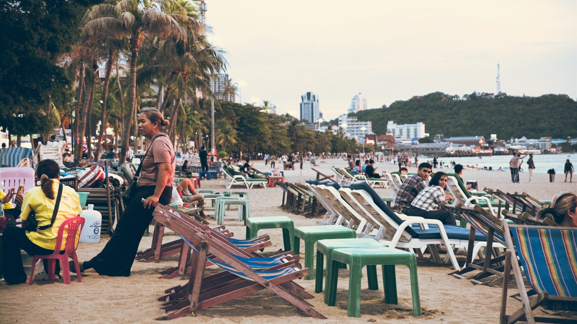 People relaxing and sitting on beach chairs and benches on a crowded sandy beach with palm trees, in front of city buildings and a hill in the background during daytime.