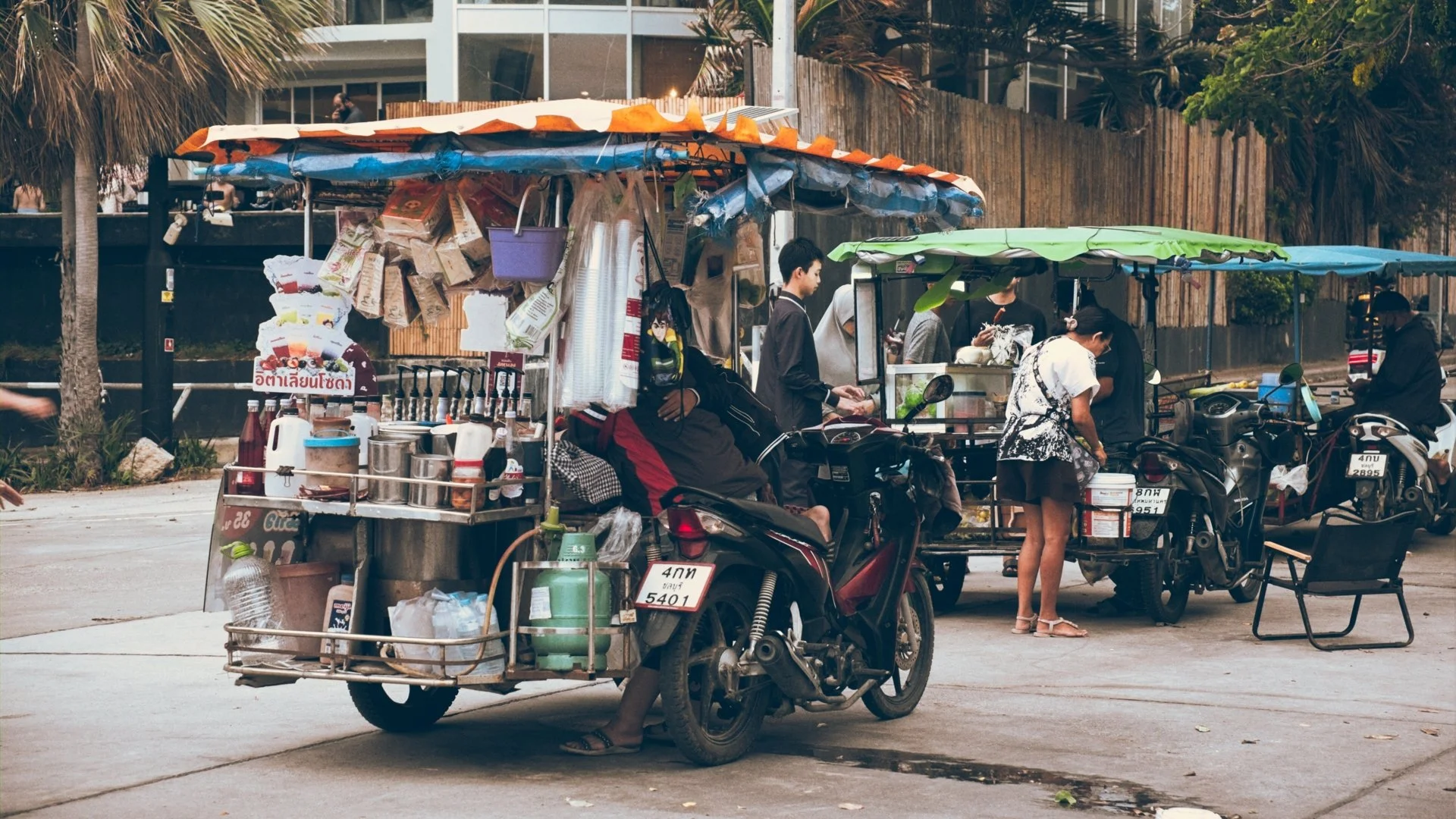 Street scene with a motorcycle cart selling snacks, with people gathered around and trees in the background.