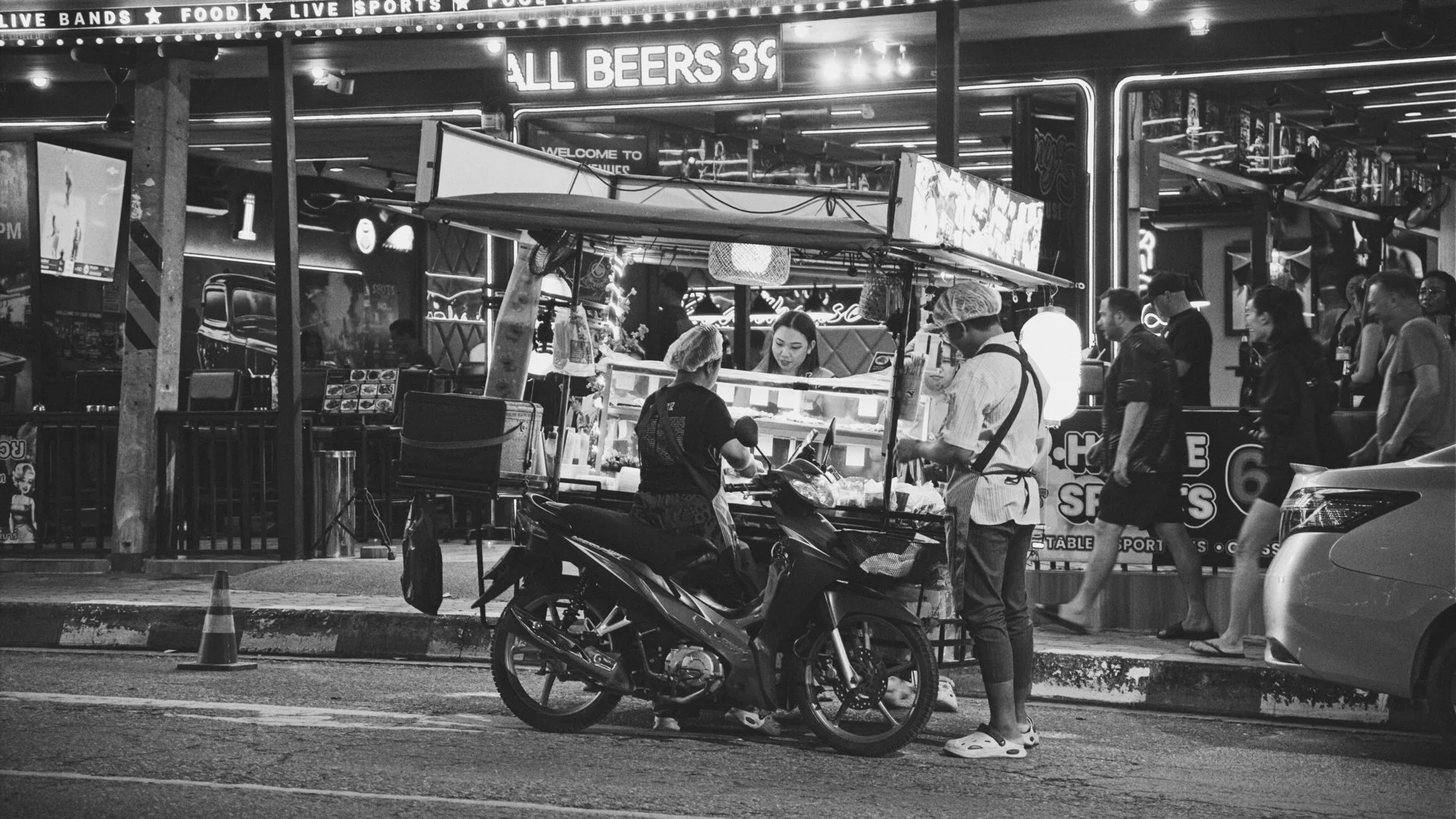 Street scene at night with a food stand, people walking, and motorcycle parked in foreground. Neon signs and lights illuminate the area.