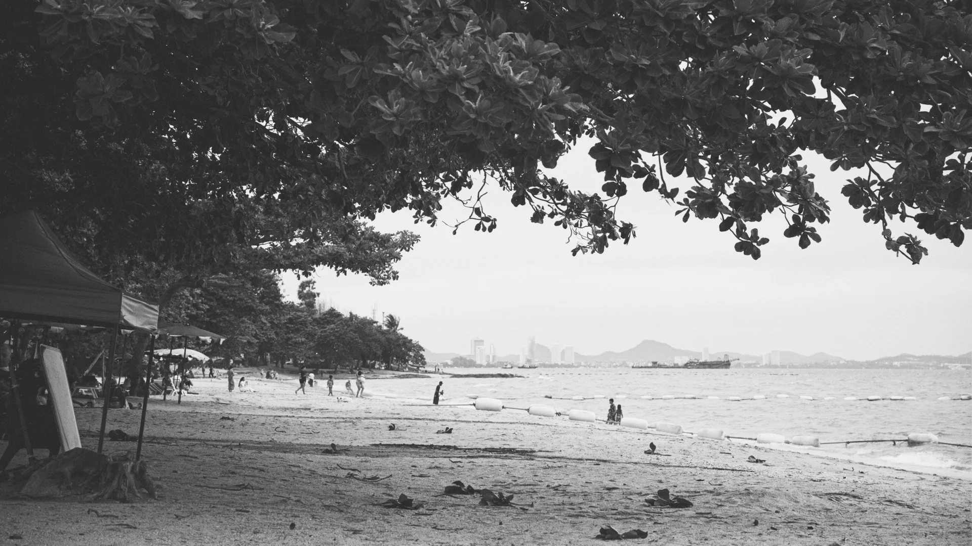 A black-and-white photo of a beach scene with trees, people walking and playing, tents, and a city skyline in the background.