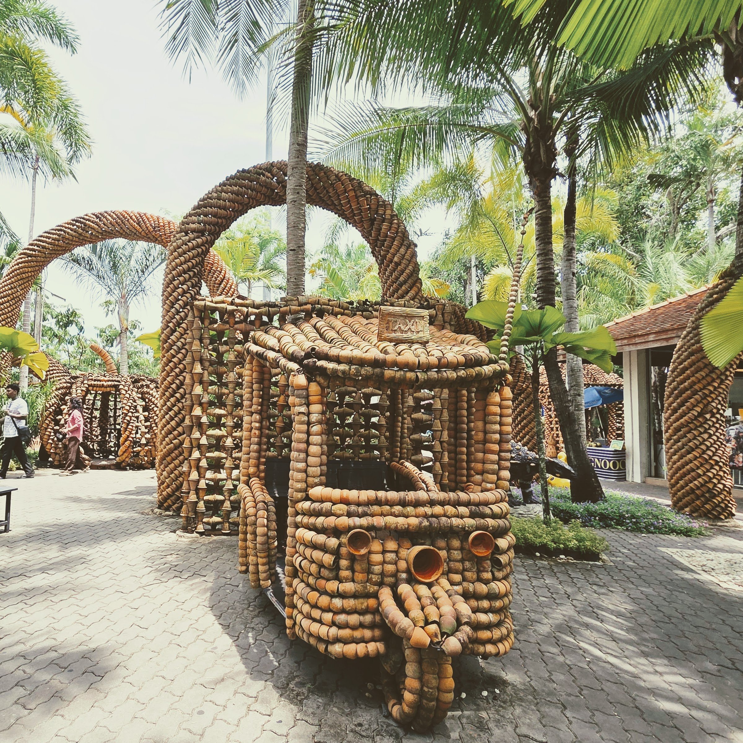 Outdoor scene with palm trees and a large decorative structure made of clay or ceramic pots arranged in spiral patterns.