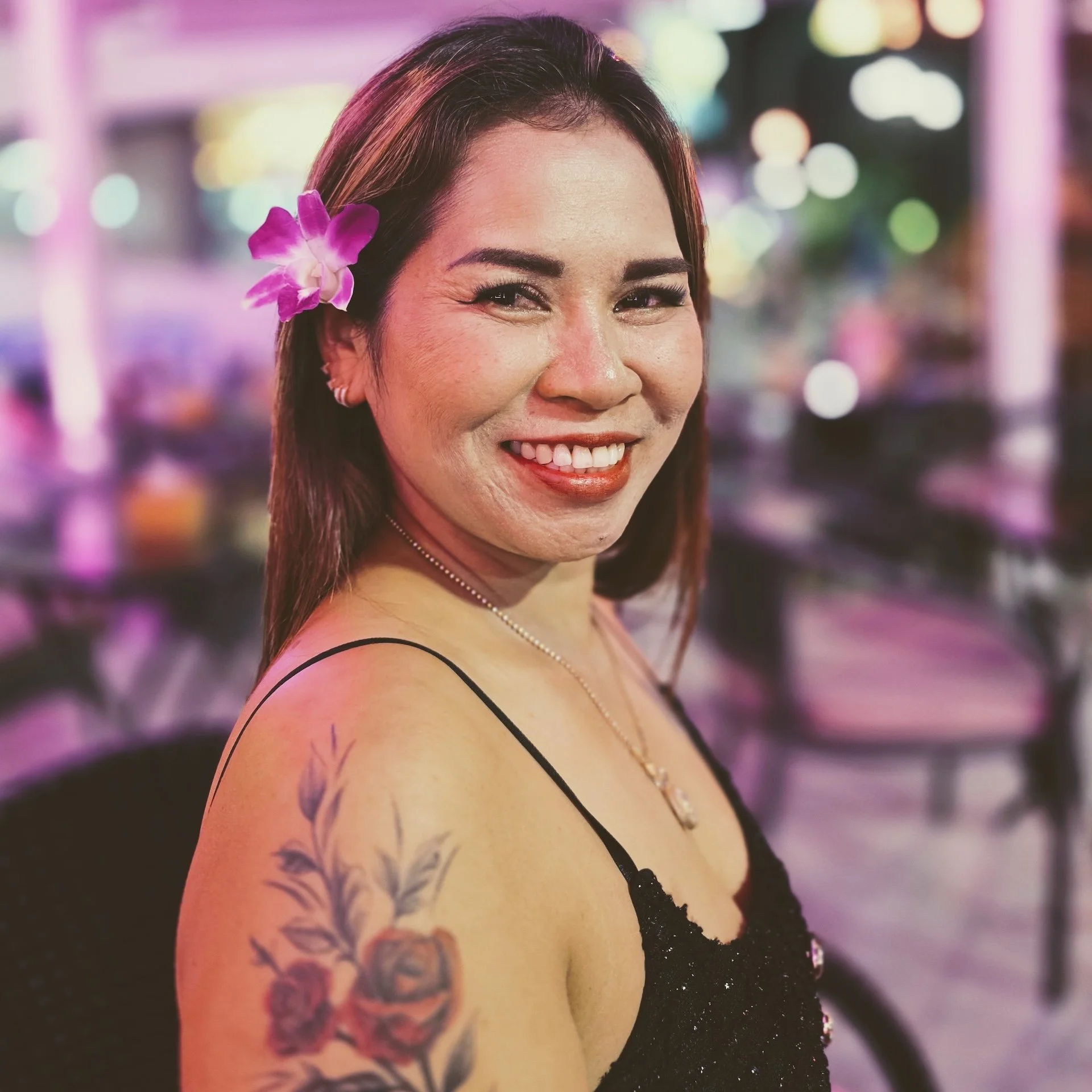 Close-up of a woman with a purple flower in her hair, wearing a black dress with thin straps, in front of a colorful, blurred city lights background.