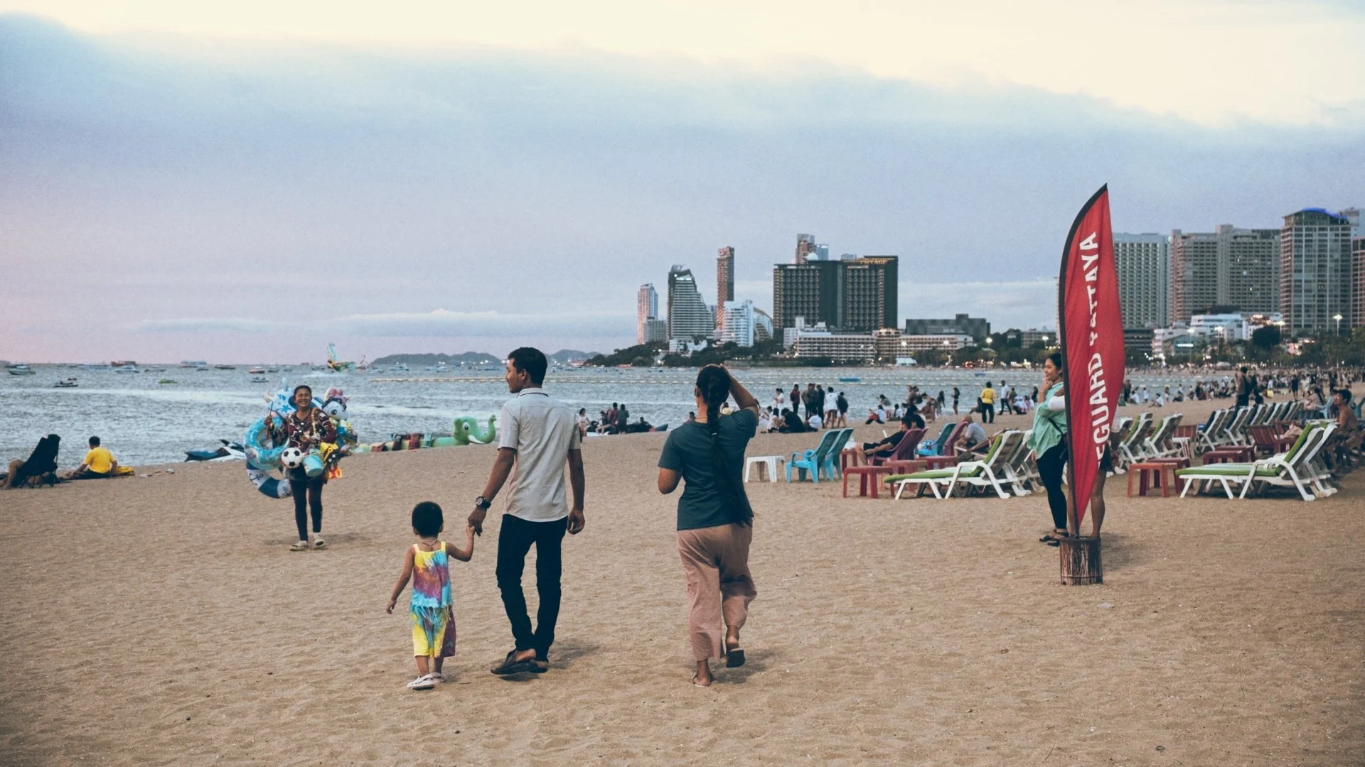 People walking on a sandy beach near the ocean with city buildings in the background, some sitting on lounge chairs, and a red banner that reads 'GUARD & VAIA'.