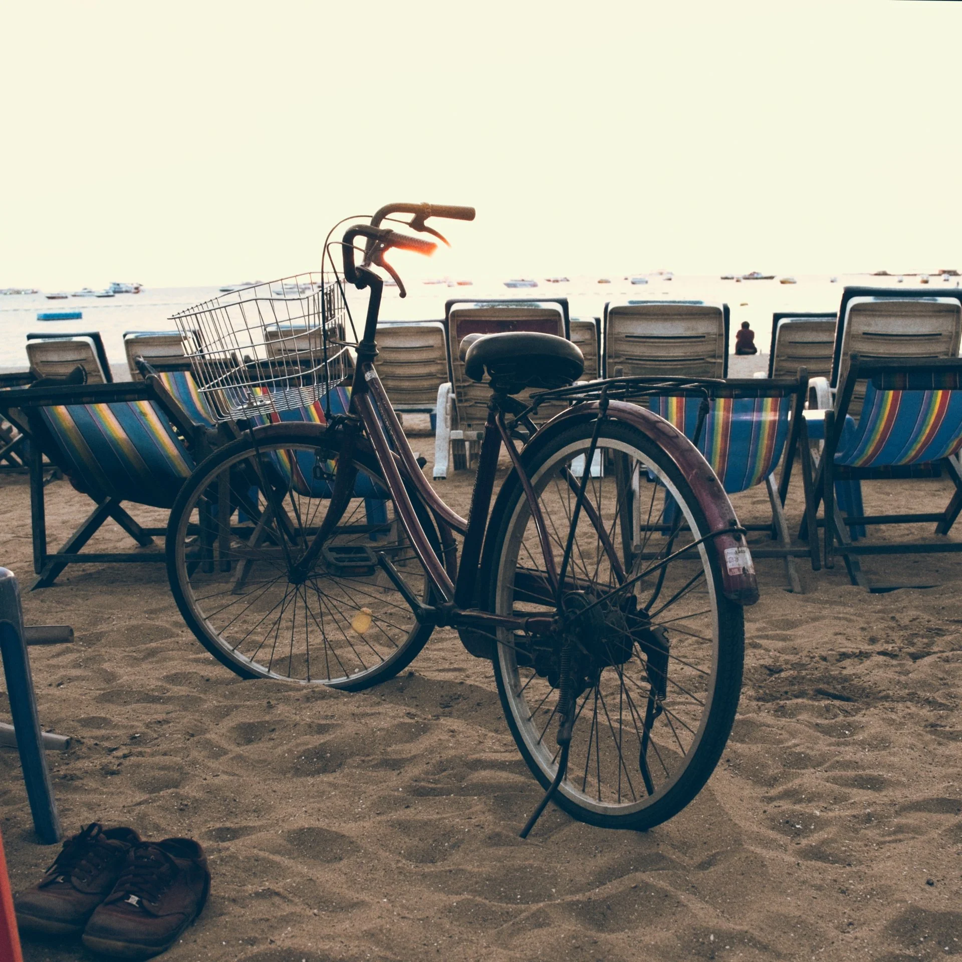 Bicycles and chairs on a sandy beach during sunset with boats in the water in the background.