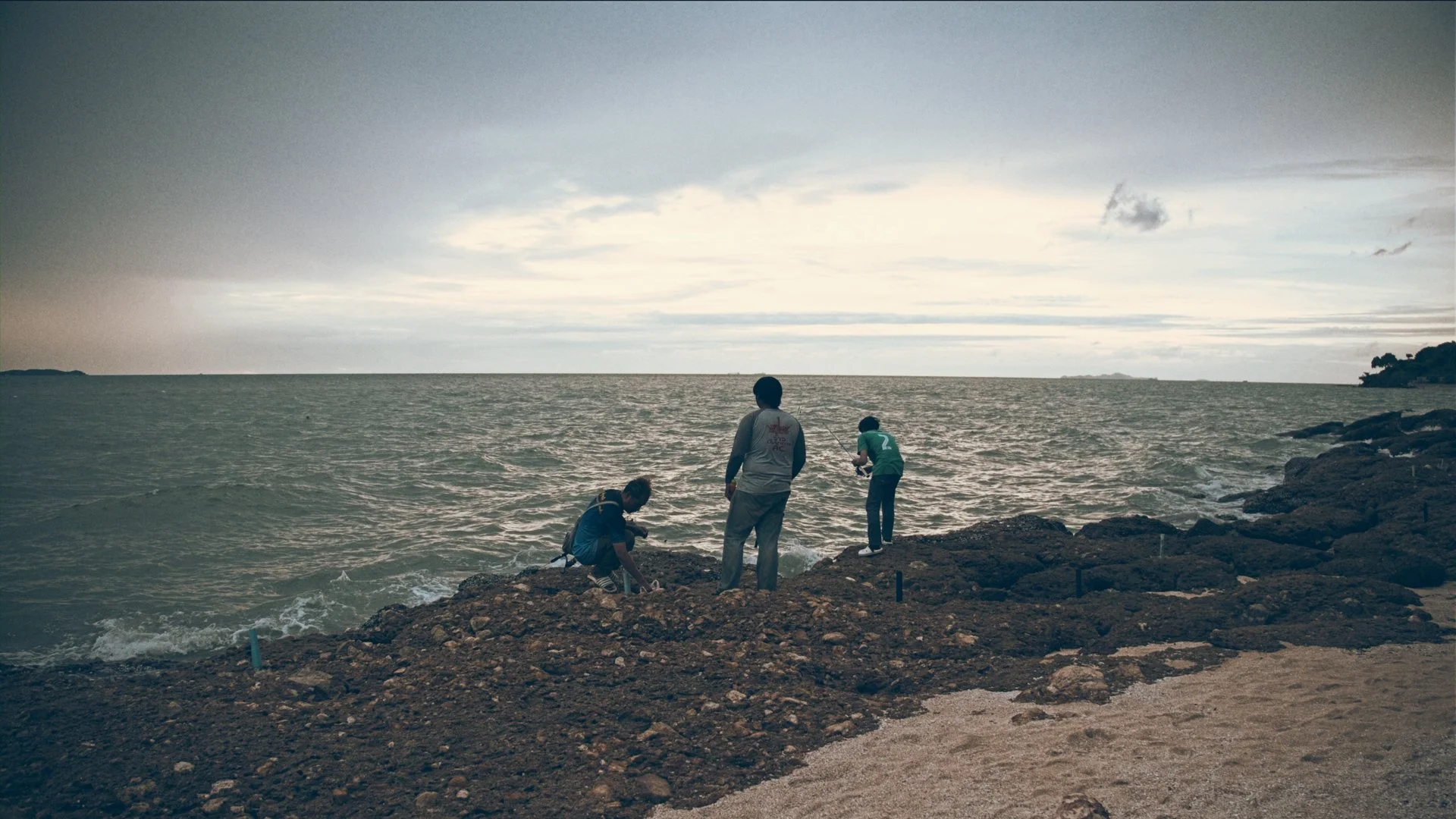 Three people standing and crouching on rocky shoreline by the ocean, with cloudy sky and distant land in the background, possibly fishing or exploring.