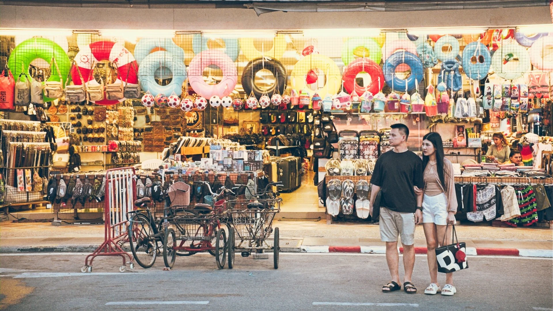 A street view of a store selling beach accessories and souvenirs, with two people holding hands walking past. The store displays colorful inflatable rings, bags, hats, beach toys, and footwear.