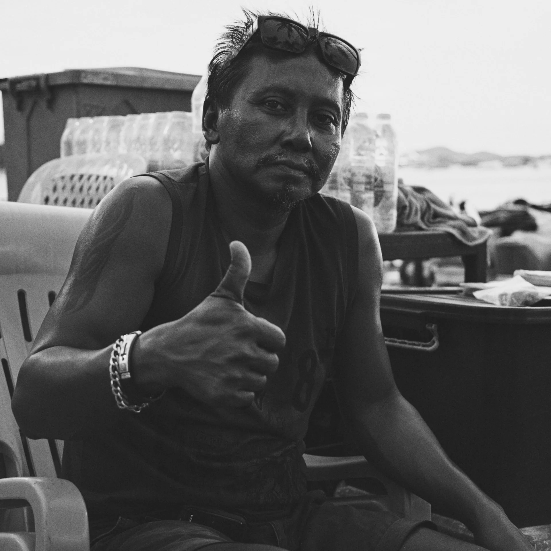 Black-and-white photo of a man with sunglasses resting on his forehead, giving a thumbs-up, sitting outdoors with water and mountains in the background.