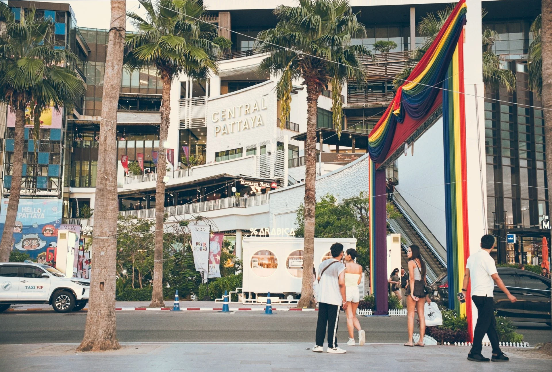 People walking in front of Central Pattaya shopping mall with tall palm trees, storefronts, and a rainbow-colored decoration structure.