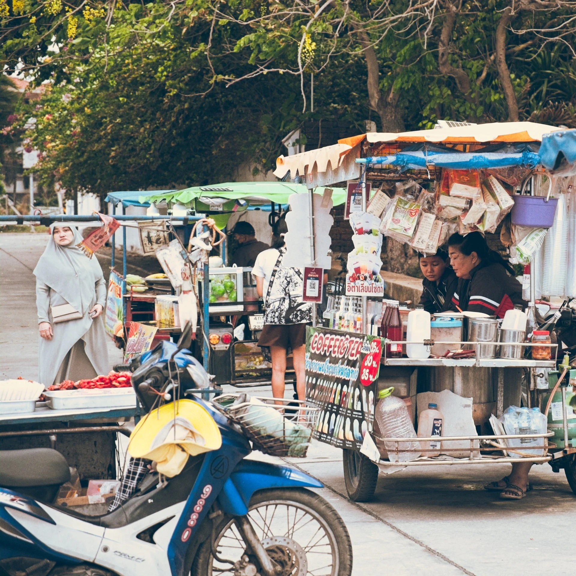 A street scene with small street food stalls covered with colorful tarps, surrounded by people, a motorcycle, and lush green trees in the background.