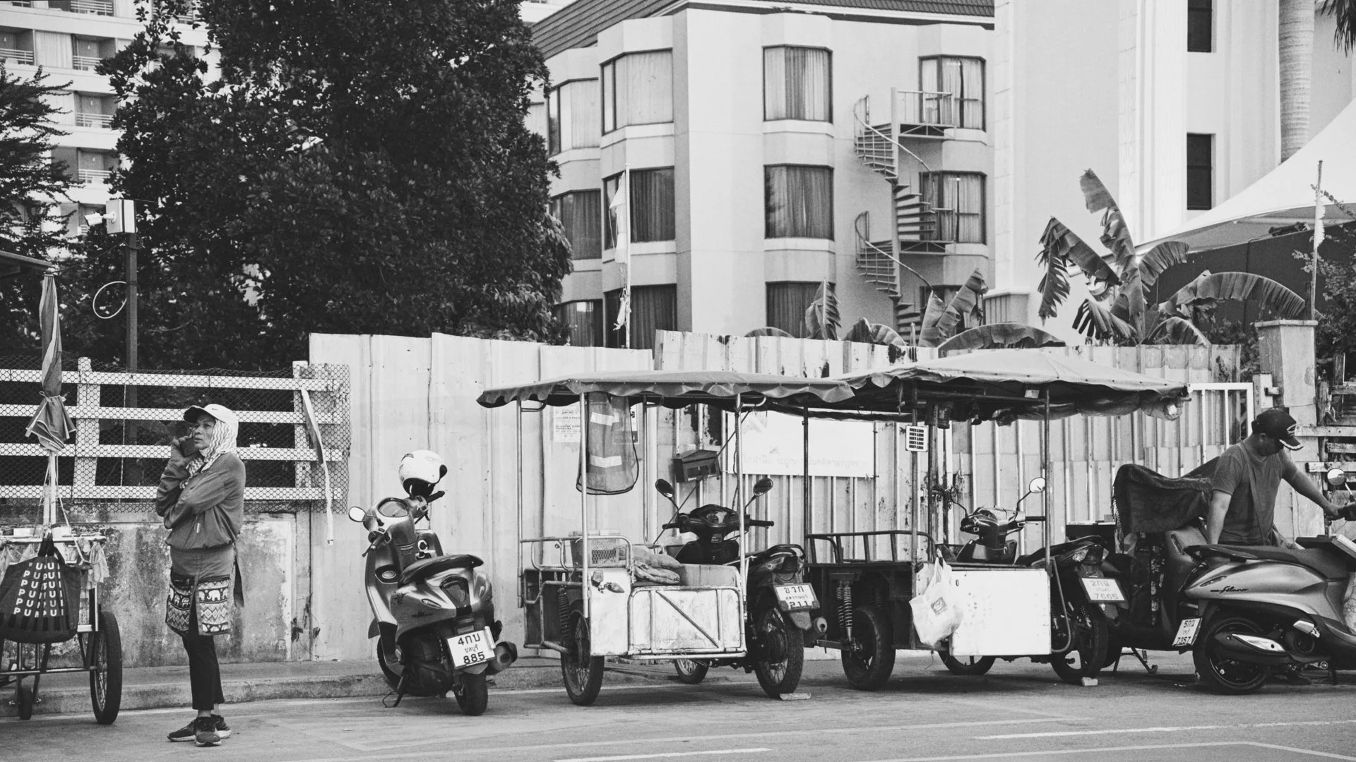 Street scene with motorcycles, food carts, and a woman with an umbrella in urban setting. Multi-story building with fire escape and large trees in the background.