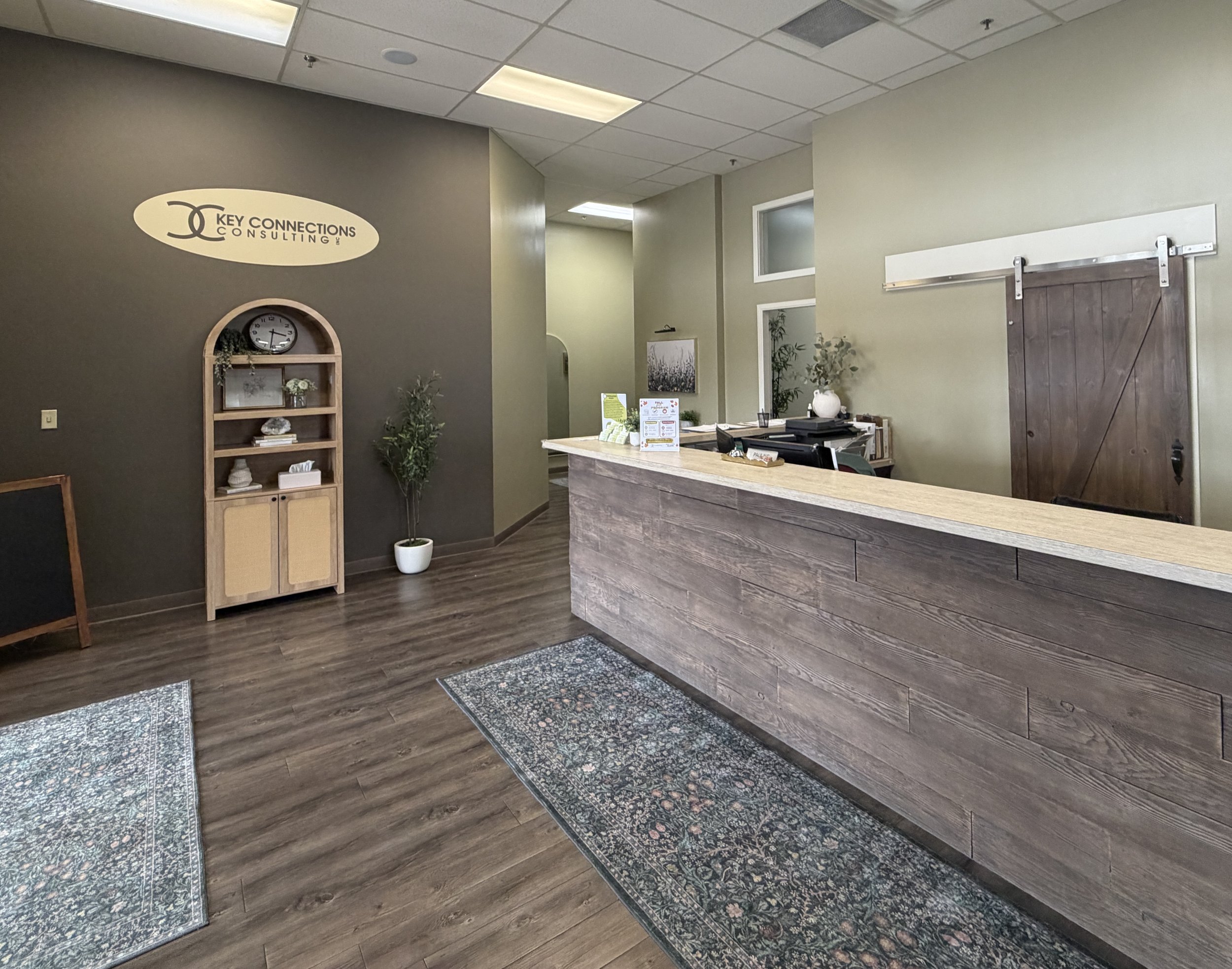 Office reception area with a wooden front desk, a wall sign reading 'Key Connections Consulting,' a bookshelf, potted plants, framed pictures, and several rugs on a wooden floor.