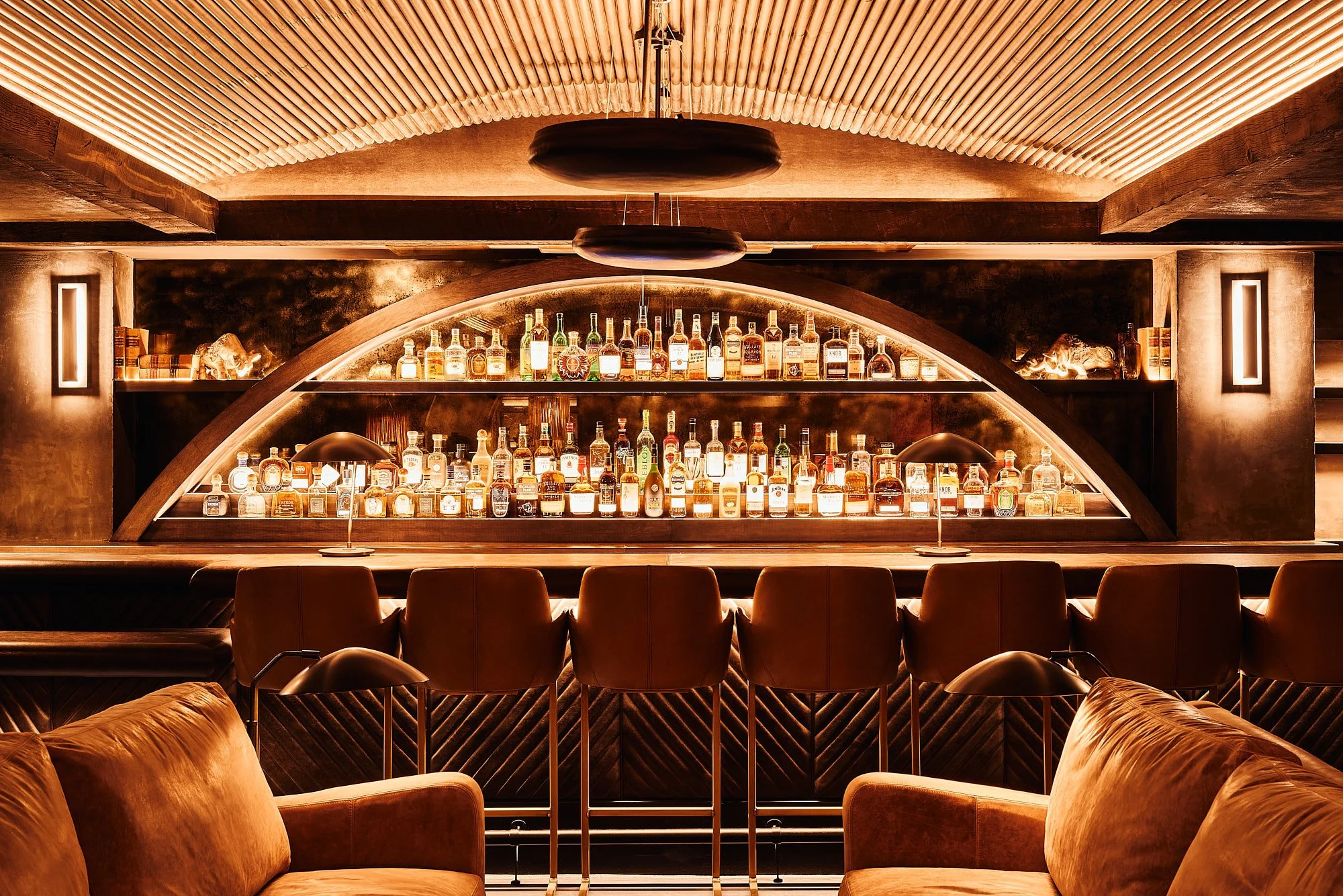 A cozy bar with warm lighting, showcasing multiple bottles of liquor on a rounded, illuminated shelf. The bar area has modern, minimalist decor with leather chairs and plush sofas in the foreground.