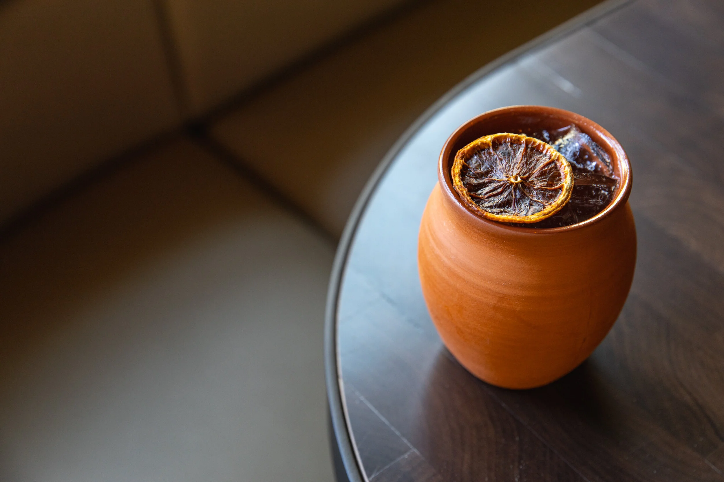 A terracotta-colored vase with a dried lemon slice on top, sitting on a dark wooden surface next to a mirror.