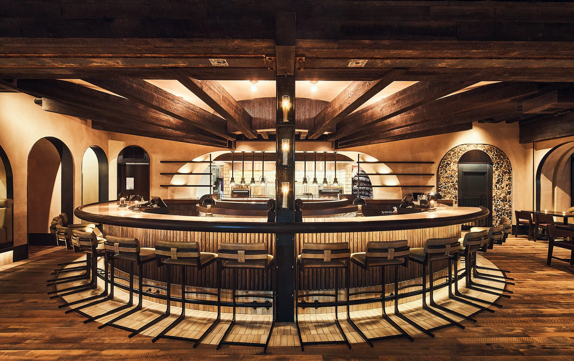 Interior of a restaurant bar with a circular bar counter, wooden beams on the ceiling, warm lighting, and surrounding chairs.