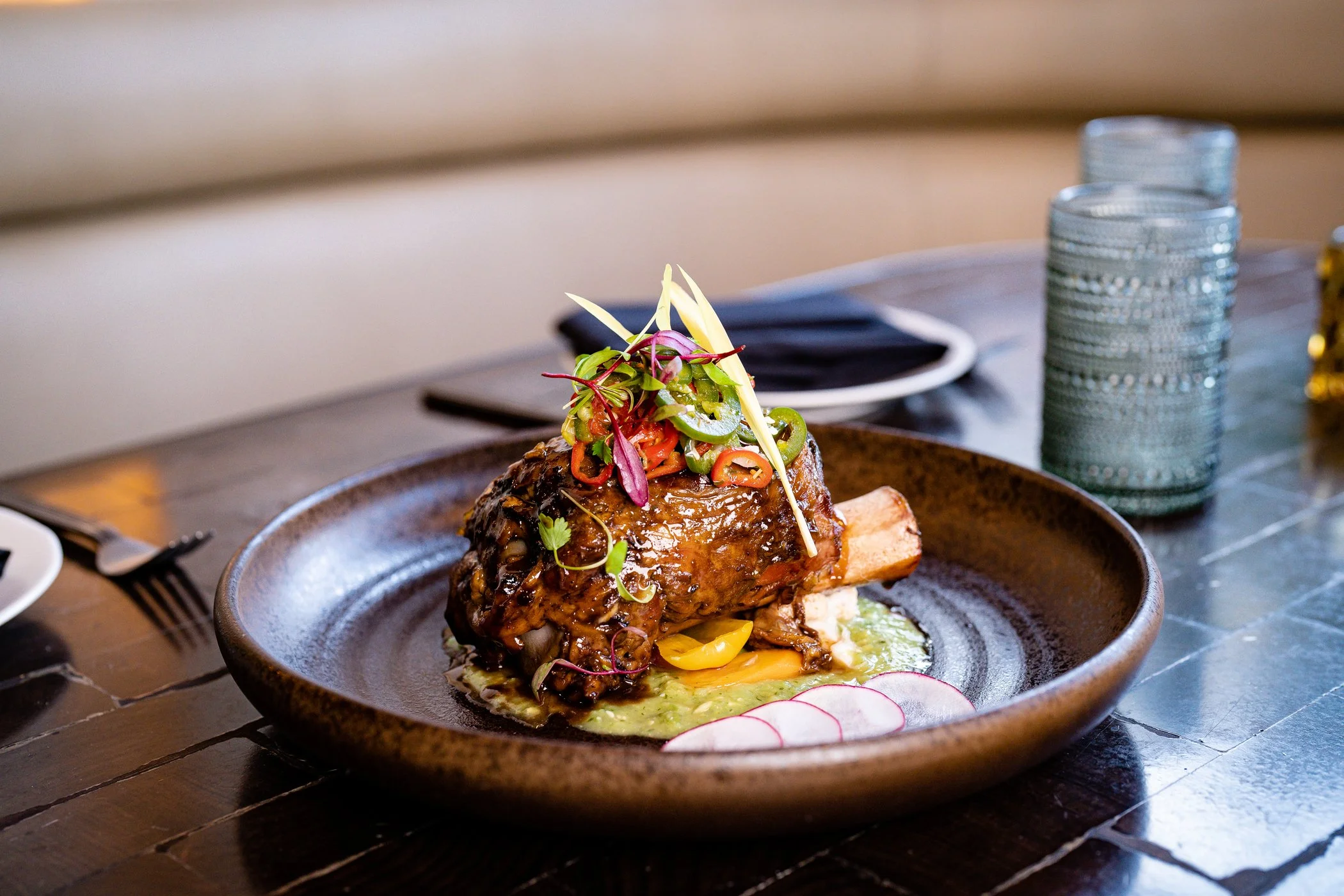 A plate of meat topped with vegetables and garnishes, placed on a dark wooden table with stools, glasses, and utensils in the background.