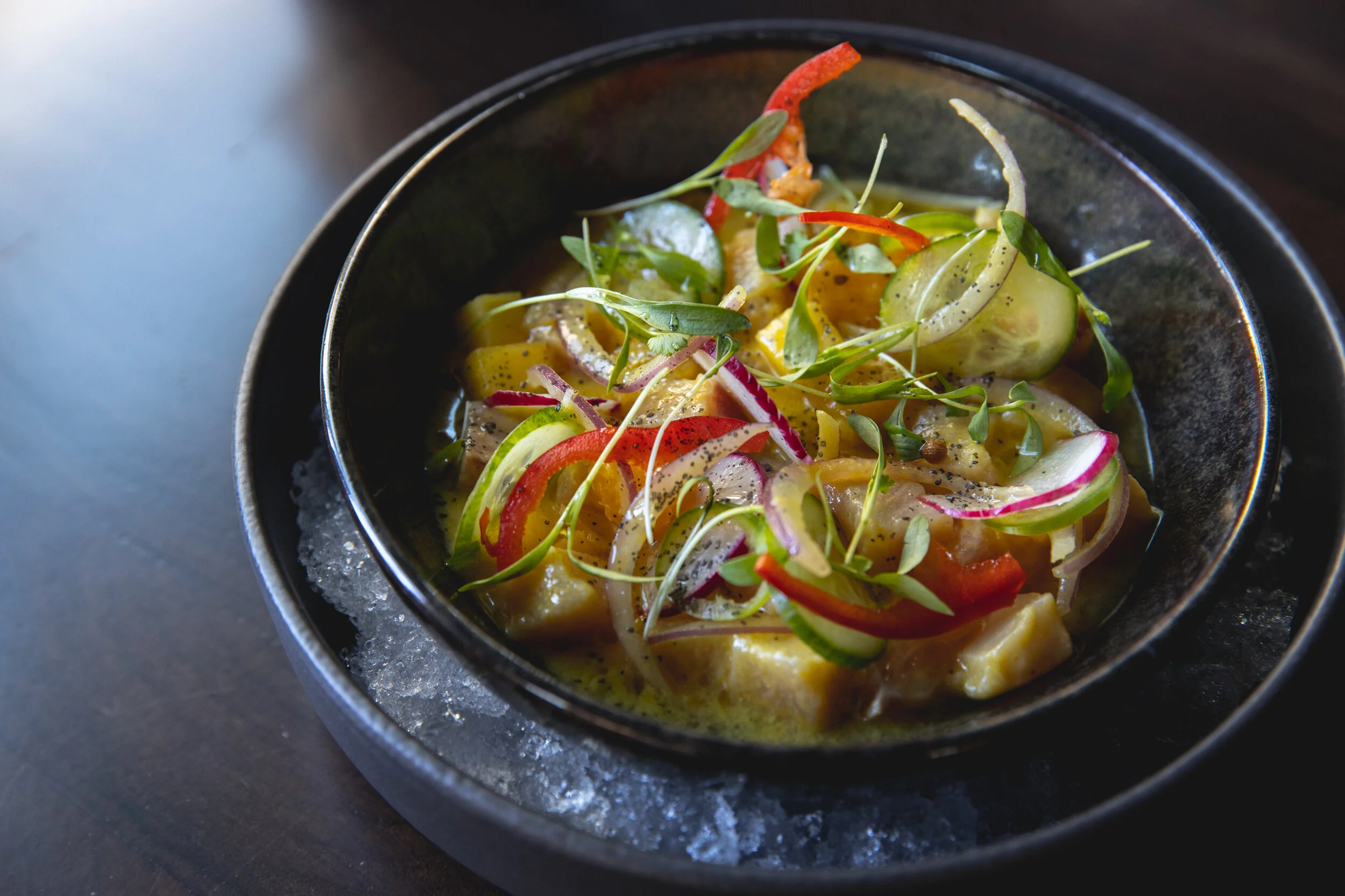 A black bowl of seafood ceviche garnished with microgreens and sliced vegetables, sitting on a bed of ice on a dark surface.