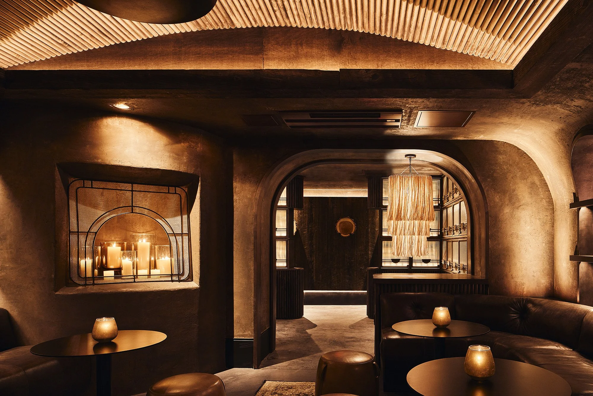 Dimly lit modern lounge with round tables, small lamps, and curved walls, featuring candles and a hanging fringed chandelier.