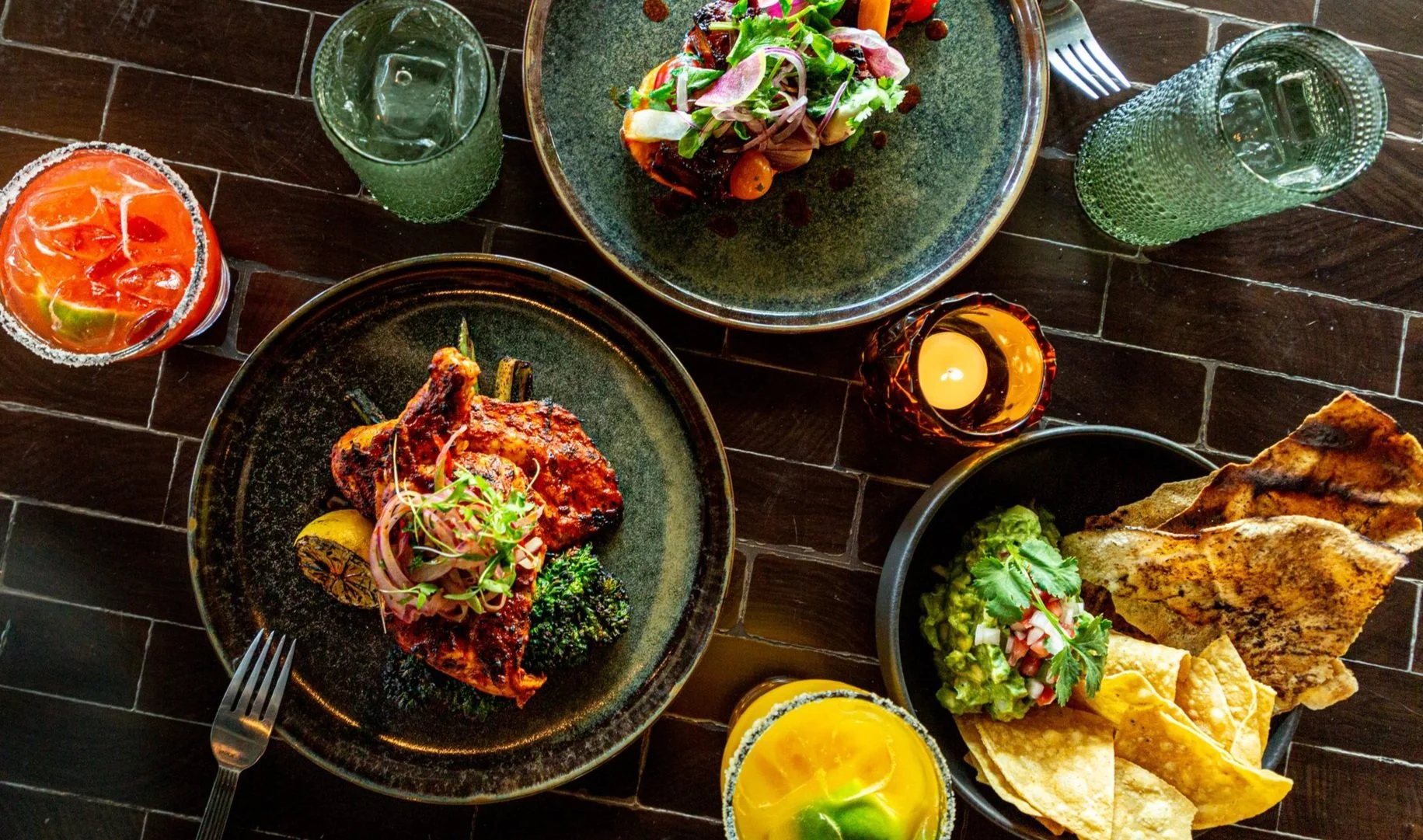 A top-down view of a dinner table with various Mexican dishes including grilled chicken, nachos, guacamole, salads, and drinks on a dark tiled surface.