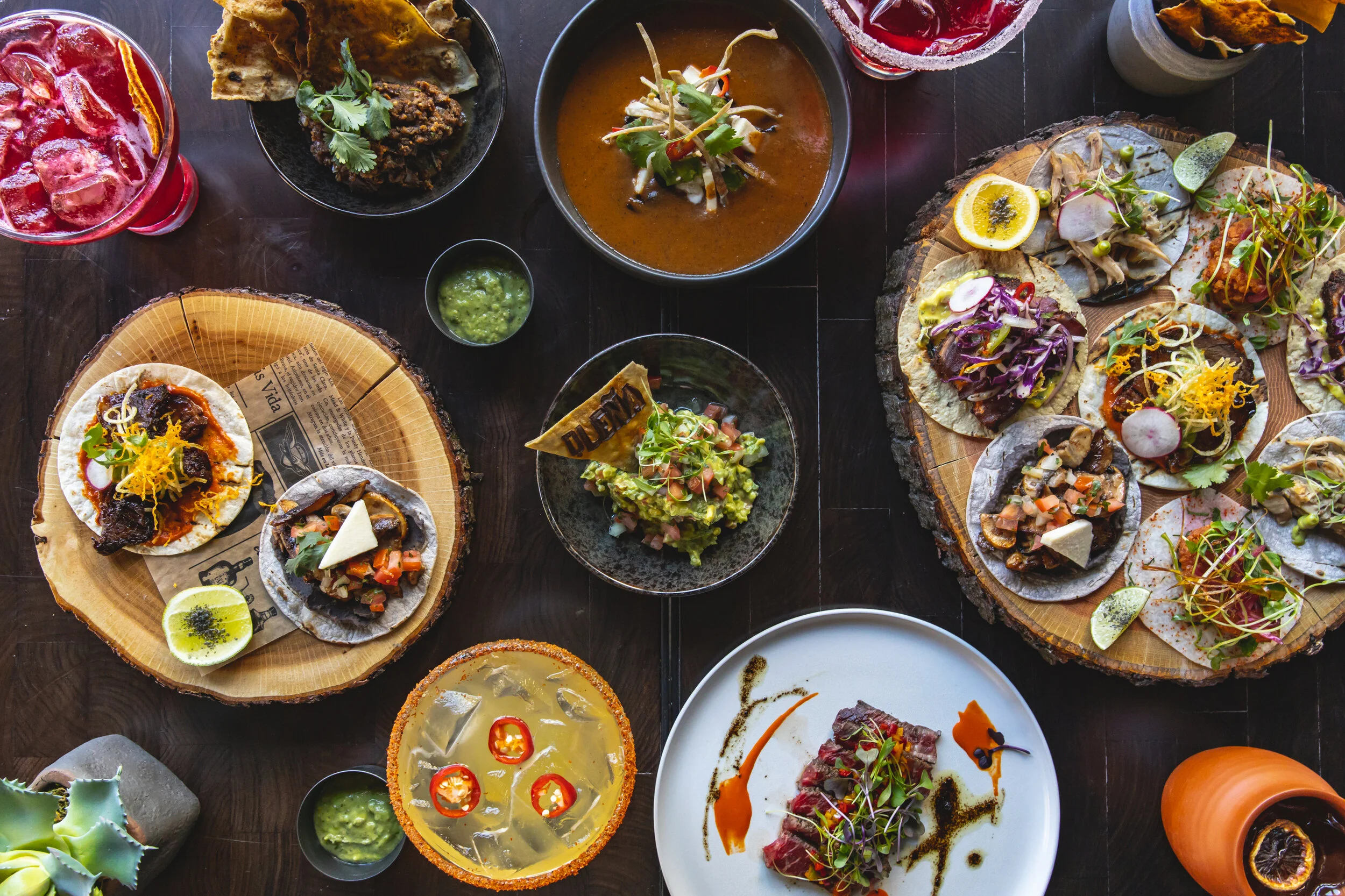 A spread of Mexican dishes including tacos with various fillings, bowls of salsa, guacamole, and soup, served on rustic wooden serving trays and small bowls on a dark table.