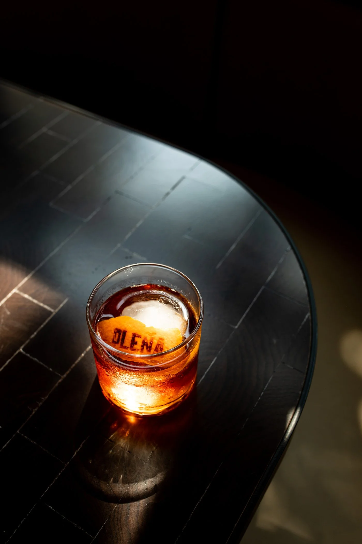 A glass of amber-colored liquid with ice and a piece of orange peel, placed on a dark round table. The orange peel has the word "BLEND" written on it. The background is black.