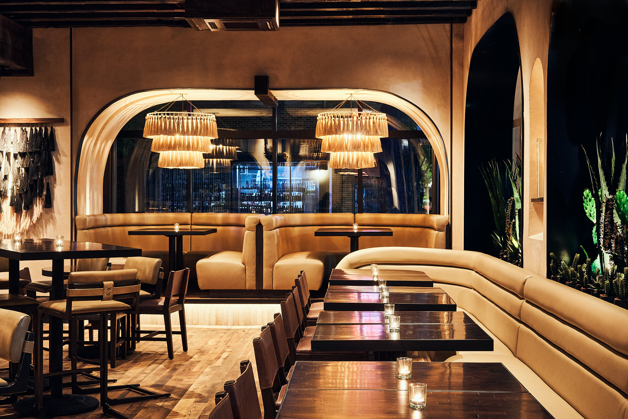 Interior of a restaurant with a warm, modern design. There are wooden tables with candles, beige curved seating along the wall, and large decorative hanging lights. Large window with a city view in the background.
