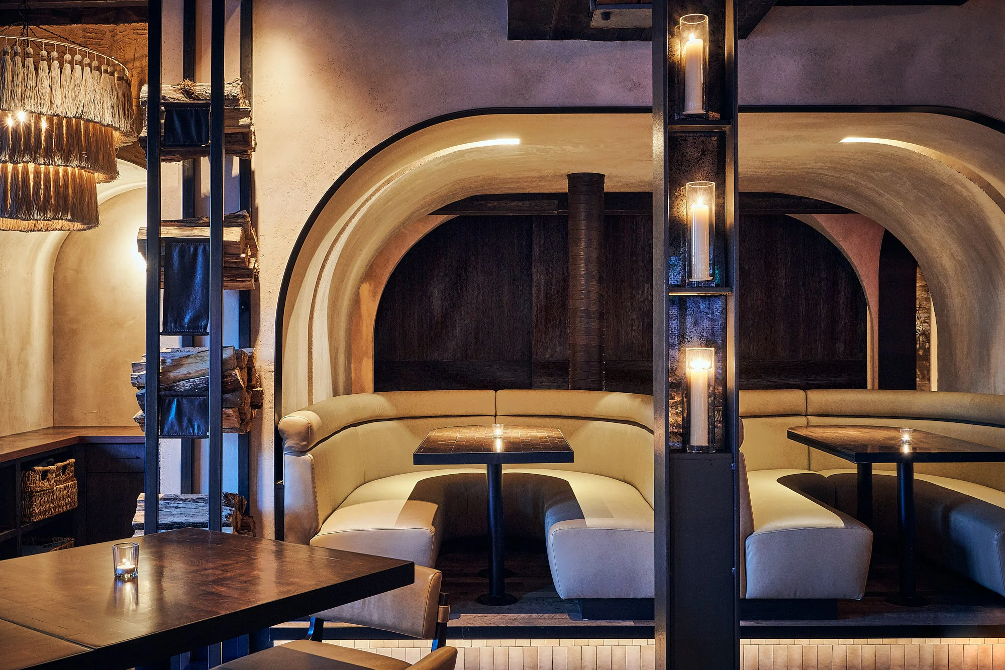 Modern restaurant interior with curved beige booths, wooden tables, candles, and decorative candles on shelves, dim lighting, and neutral colors.