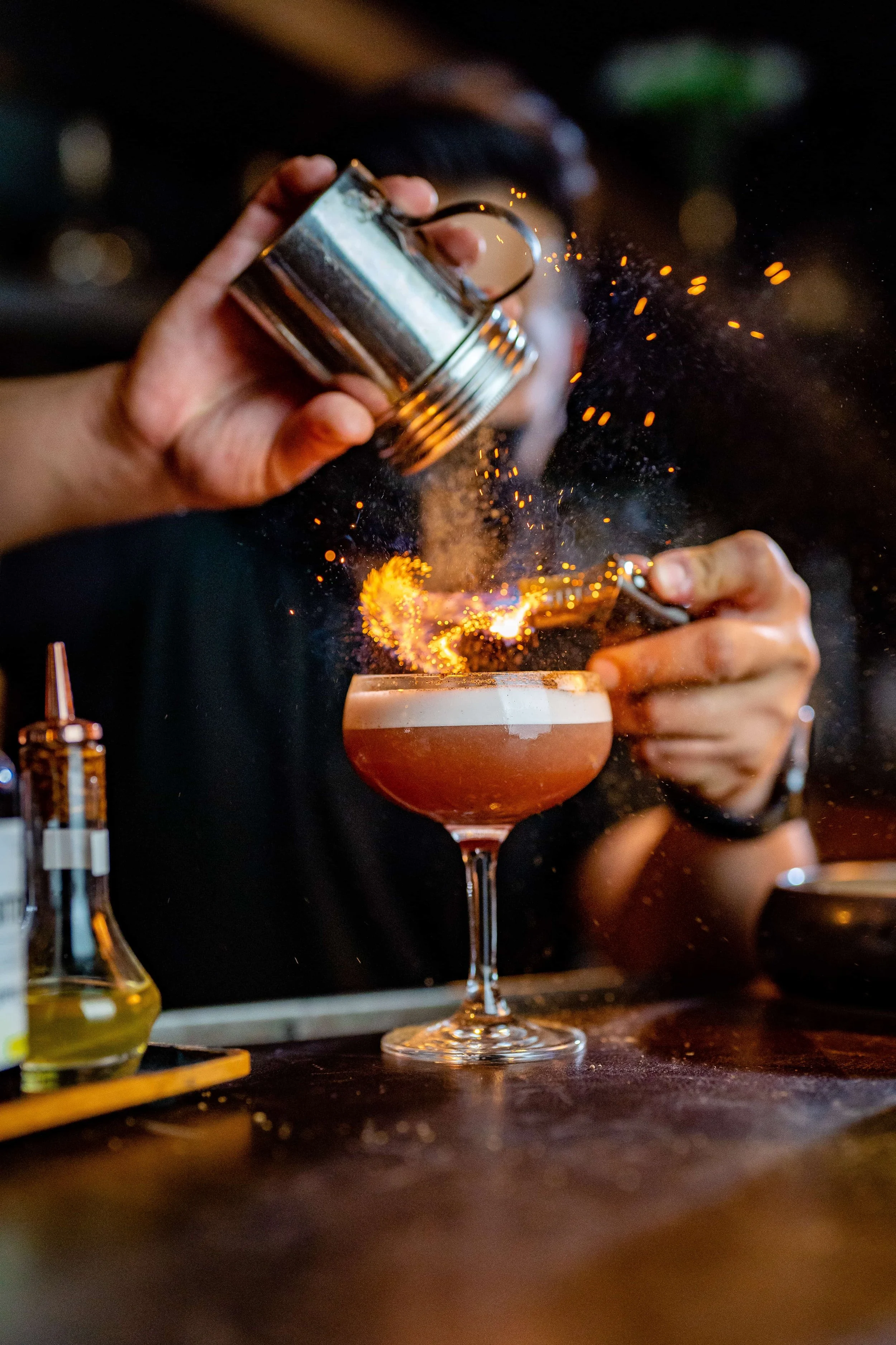 Person holding a metal shaker pouring a drink into a cocktail glass with a straw and foam topping, with sparks and a small flame in the background.