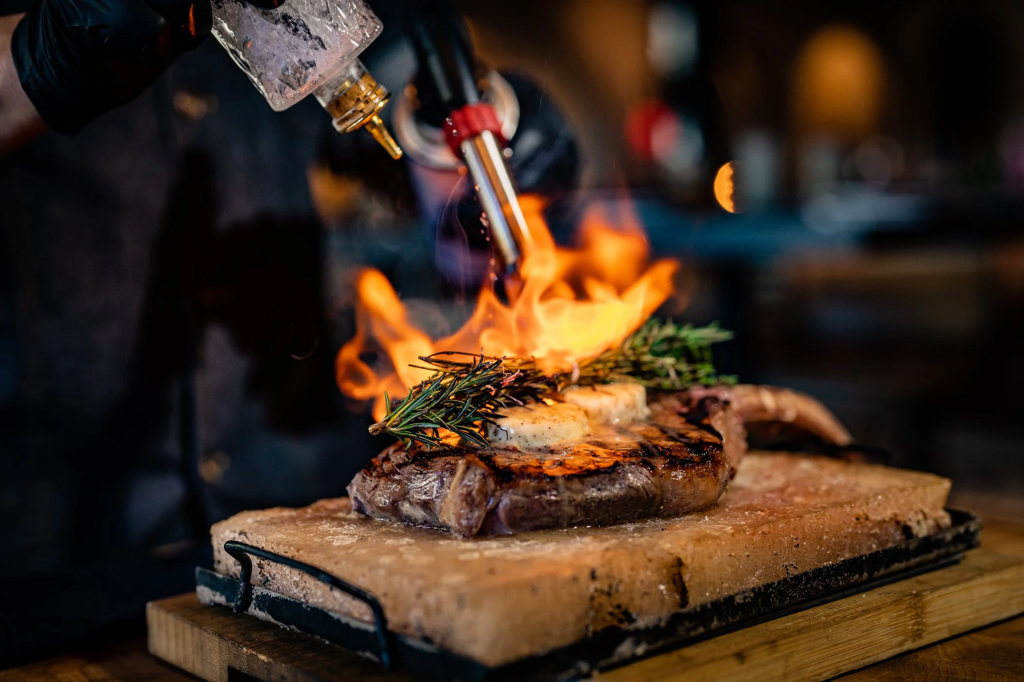 A steak topped with cheese and rosemary, being grilled with open flames from a blowtorch.