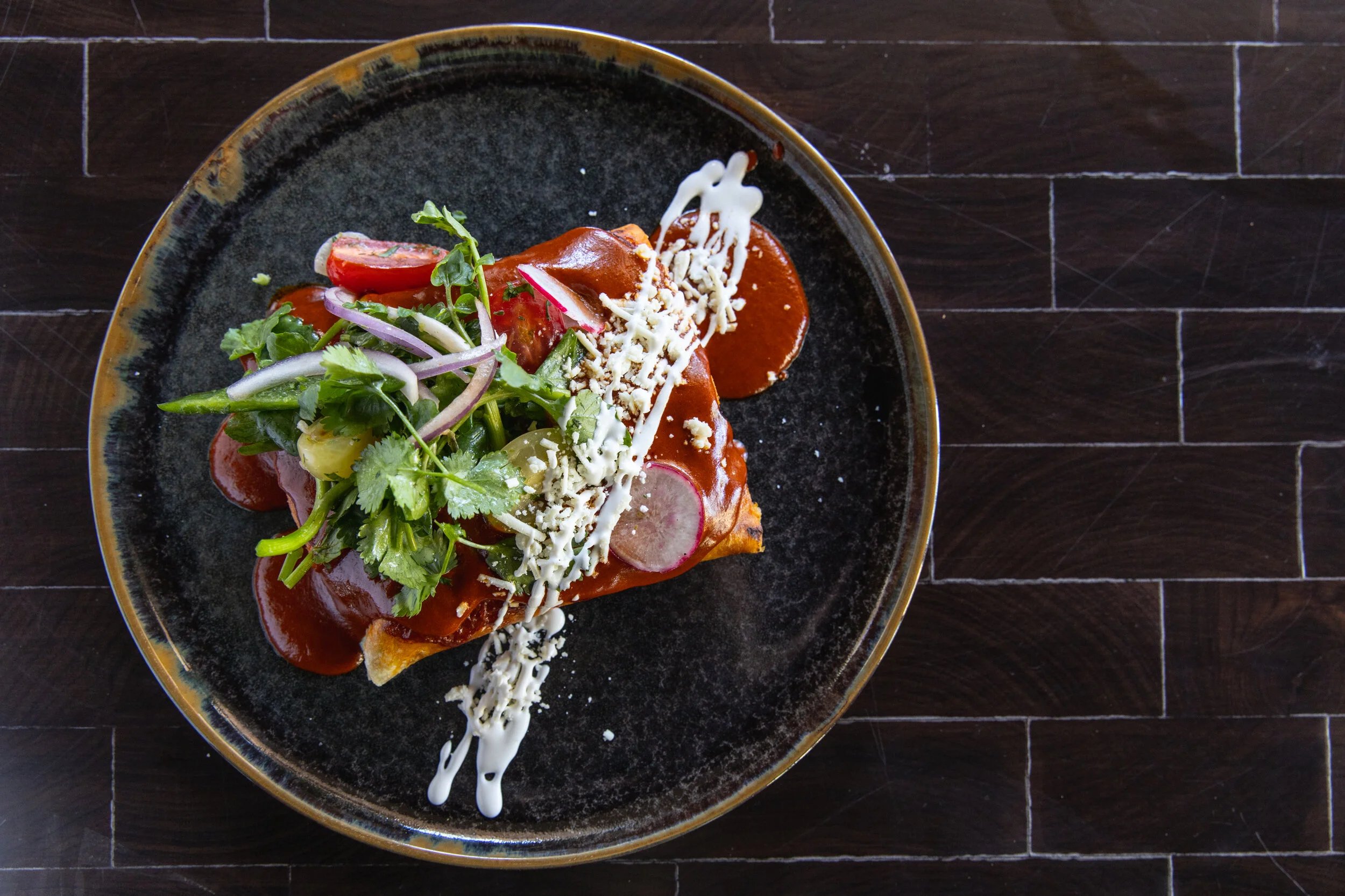 A plate of Mexican food with a tortilla, topped with red sauce, shredded cheese, sour cream, and garnished with fresh salad greens, cherry tomato halves, radish slices, and cilantro leaves on a dark wooden table.