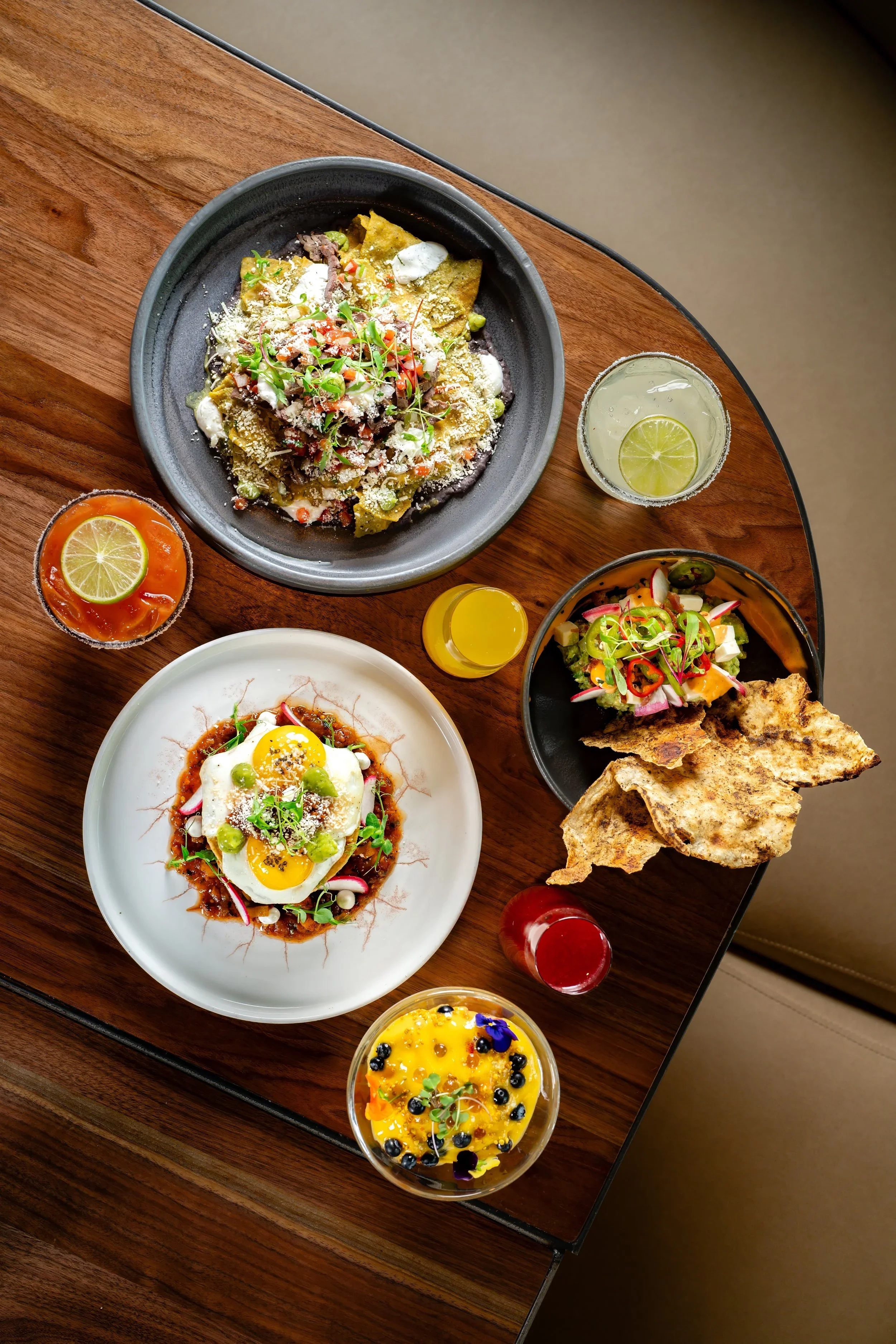 A wooden table with various dishes including a plate of nachos with cheese, sour cream, and microgreens, a fried egg on a plate with sauce and microgreens, a salad with radish, cucumber, and peppers, and a dessert with blueberries and edible flowers, along with drinks such as a lime soda, a yellow juice, and a fruit-topped dessert with blueberries.
