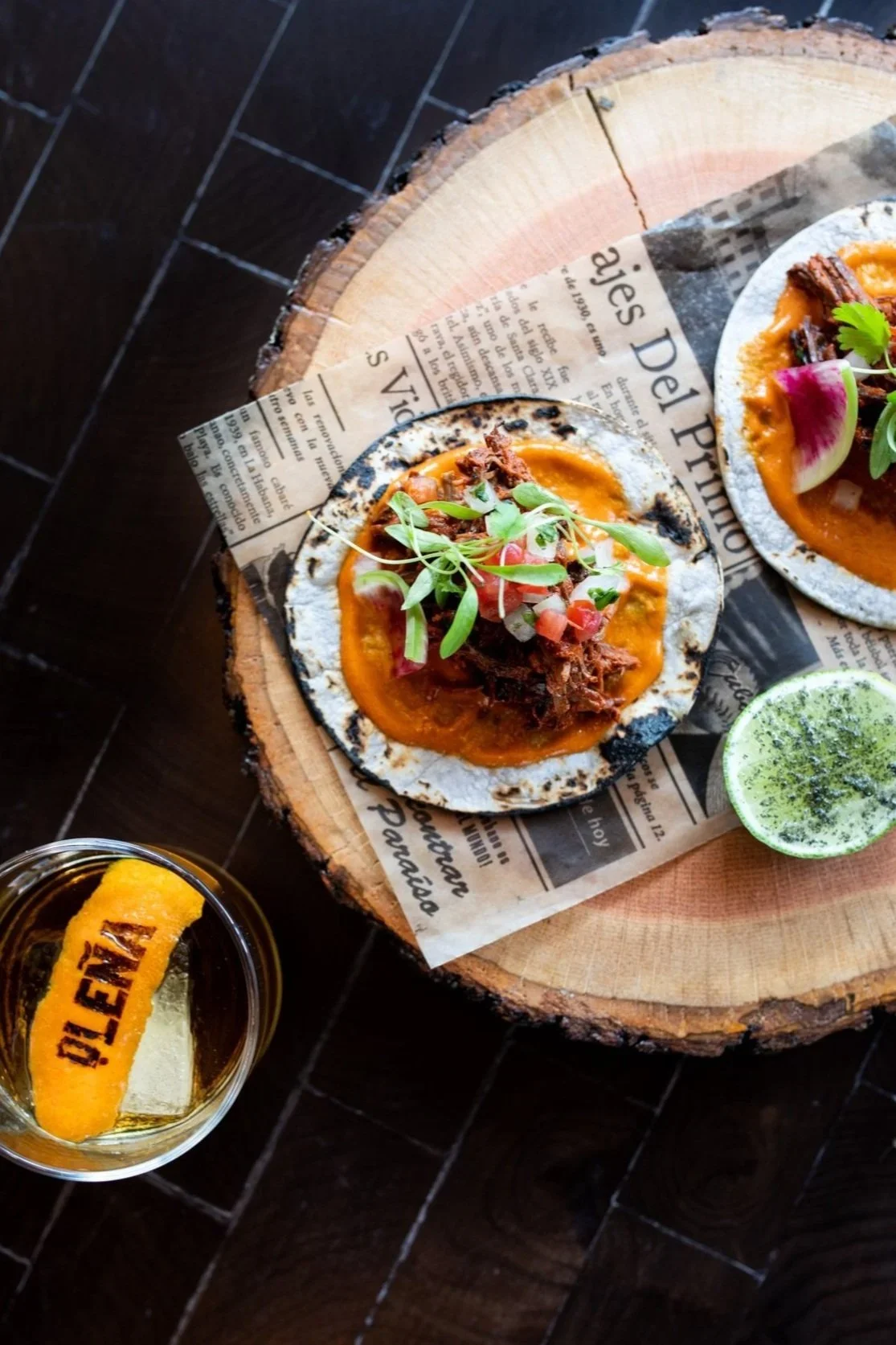 Close-up of three Mexican tacos topped with meat, herbs, and salsa, served on a wooden plate lined with newspaper-style paper, with a lime wedge and a glass of beer with an orange garnish nearby.
