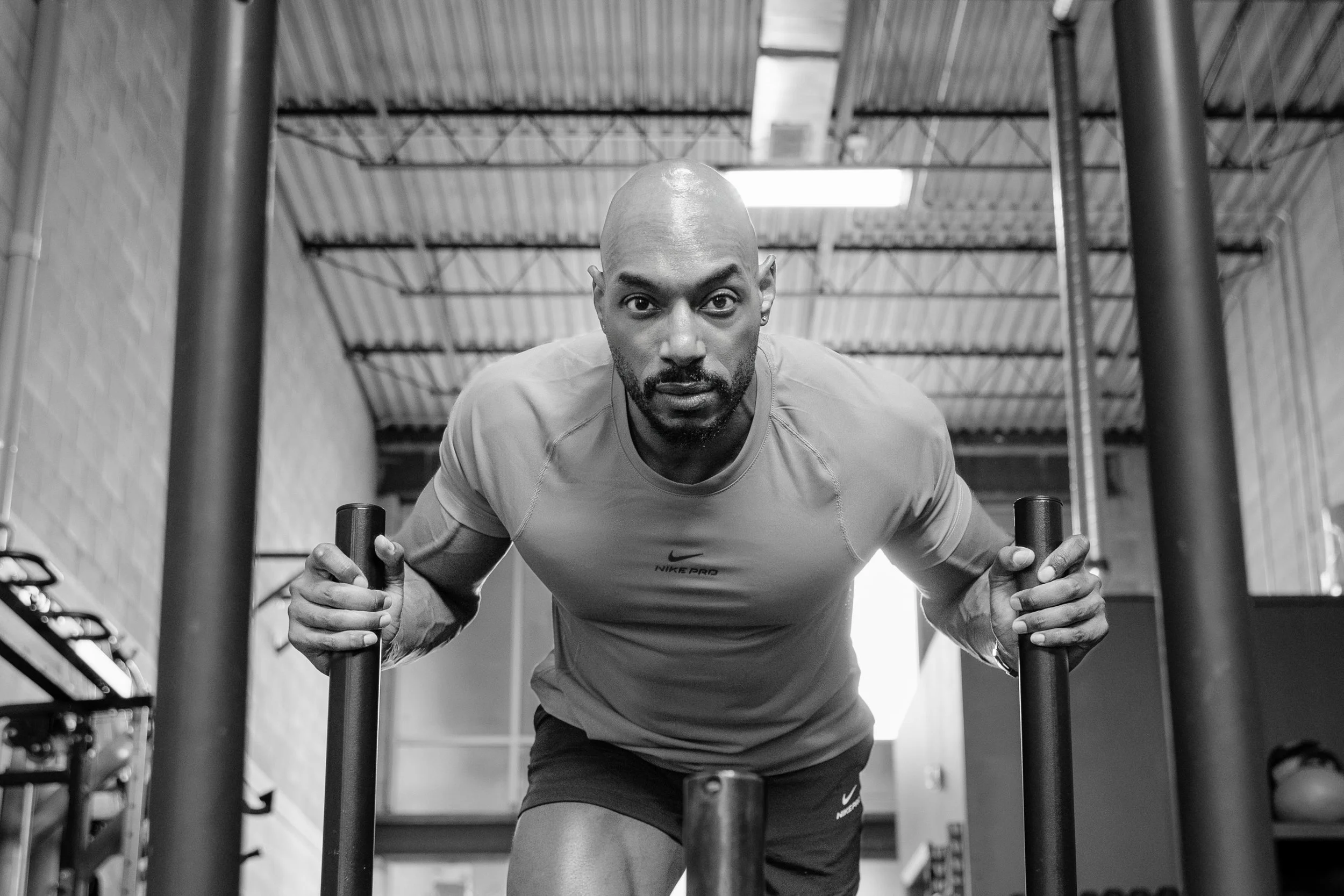 A man in a gym push-up position, holding onto workout bars, looking directly at the camera, in black and white.