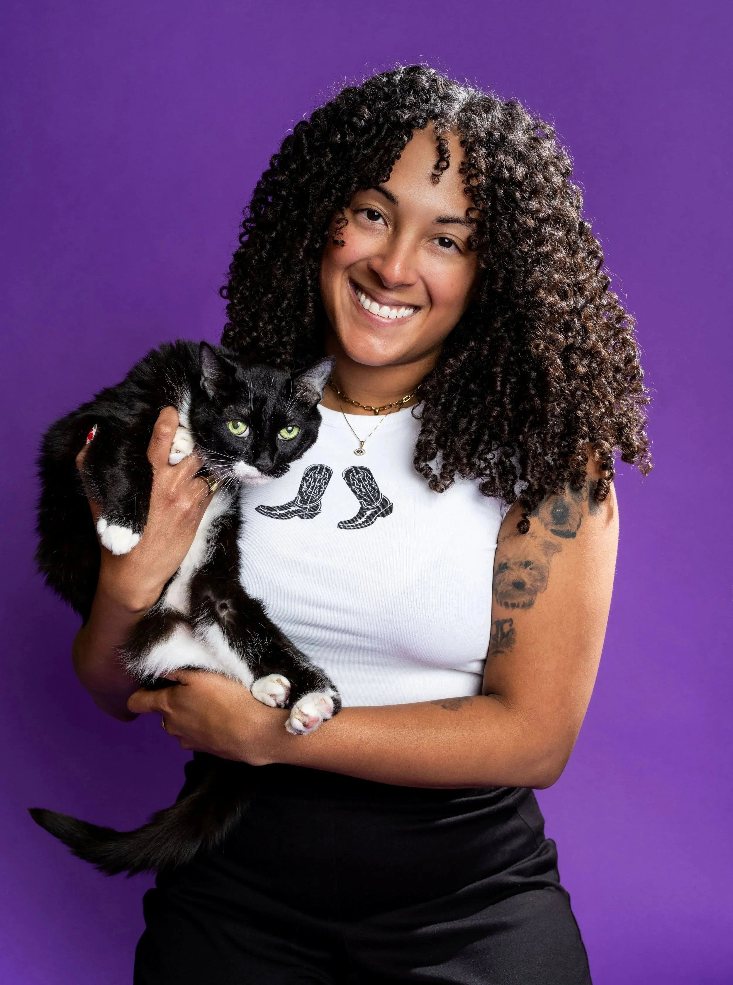A woman with curly hair smiling and holding a black and white cat, against a purple background.