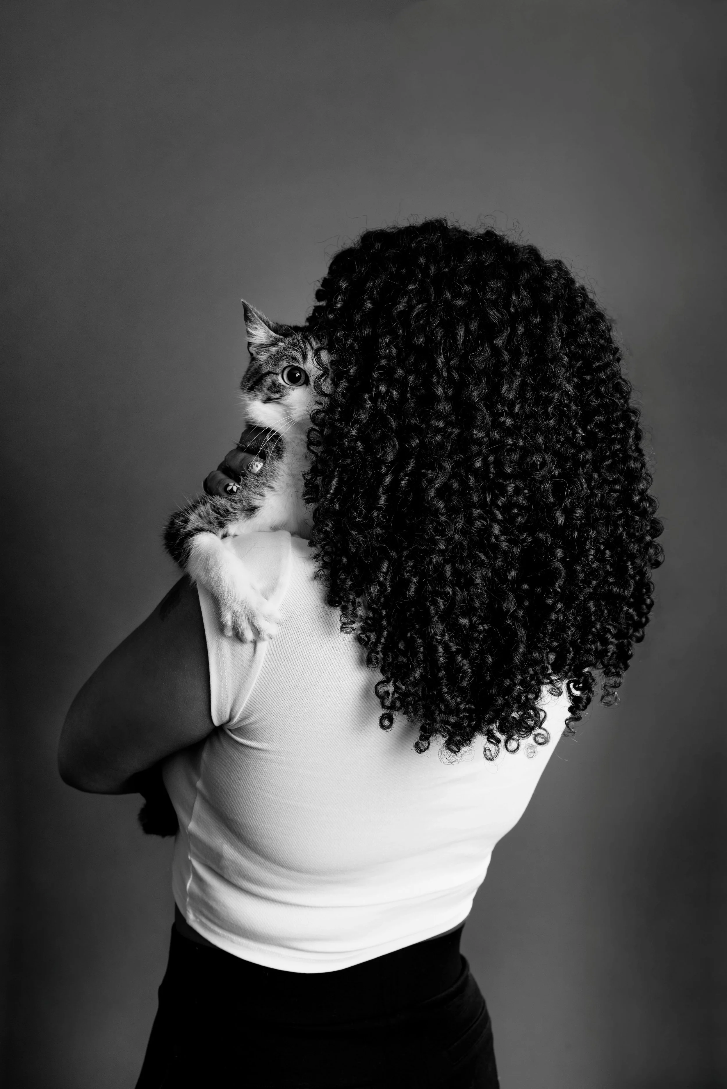 A woman with long, curly hair is holding a cat close to her face and looking at the camera, wearing a white short-sleeve shirt against a plain background.