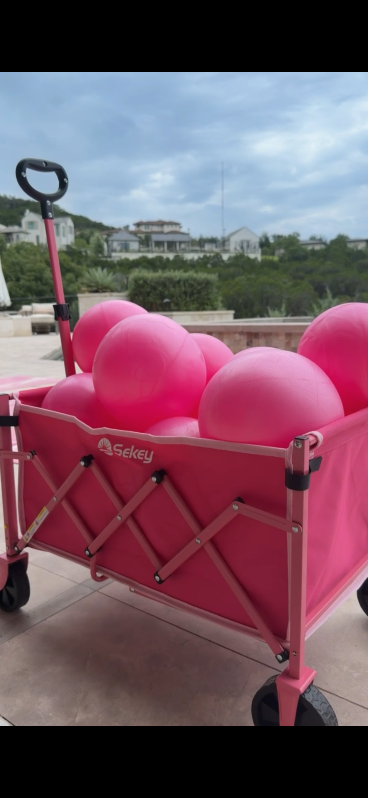 Pink wagon filled with large pink exercise balls outdoors on a patio, with houses and cloudy sky in the background.