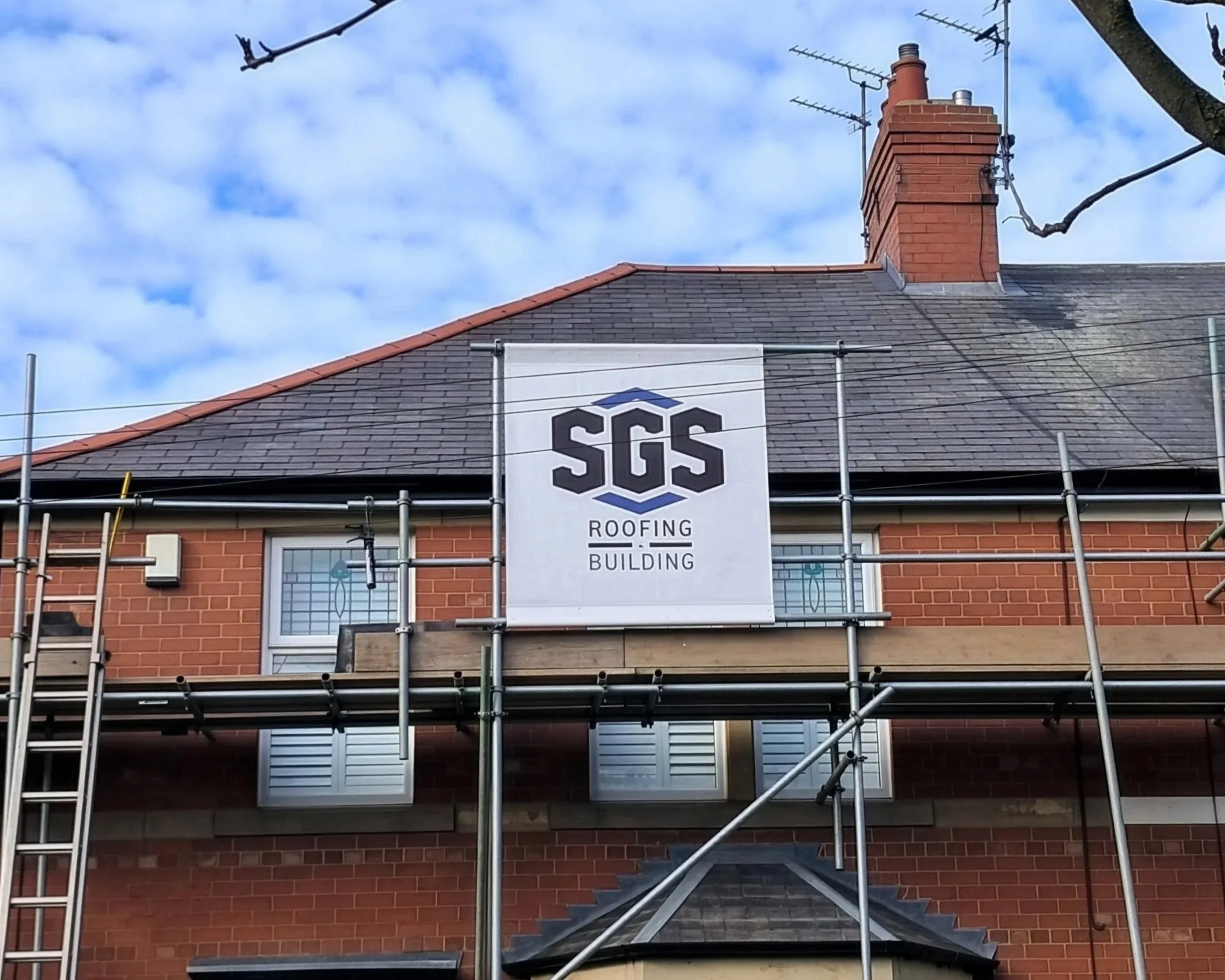 Photograph of a building under construction with scaffolding. There is a sign that reads 'SGS Roofing Building' hanging on the scaffolding. The roof is dark grey with red brick walls. A chimney and multiple antennas are visible on the roof, and a large window with white blinds is in the bottom part of the building. The sky is partly cloudy.