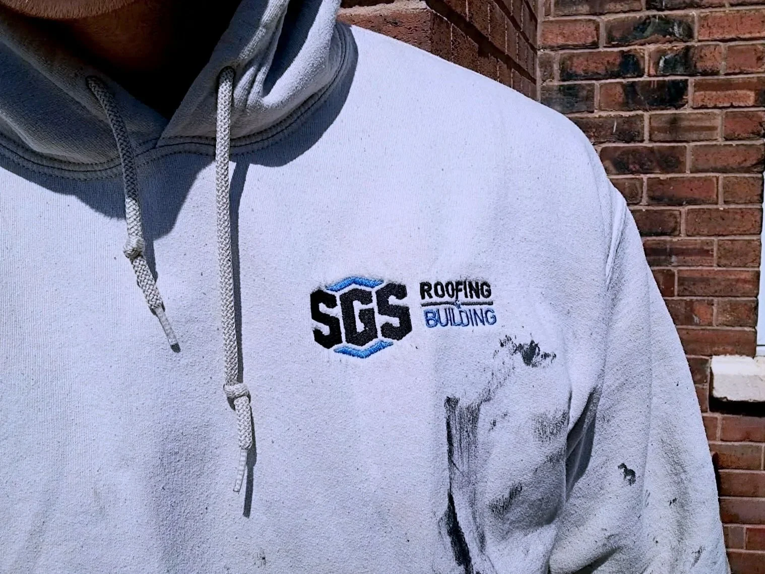 A person wearing a light gray hoodie with an embroidered logo reading 'SGS Roofing Building' on the chest, standing in front of a red brick wall.