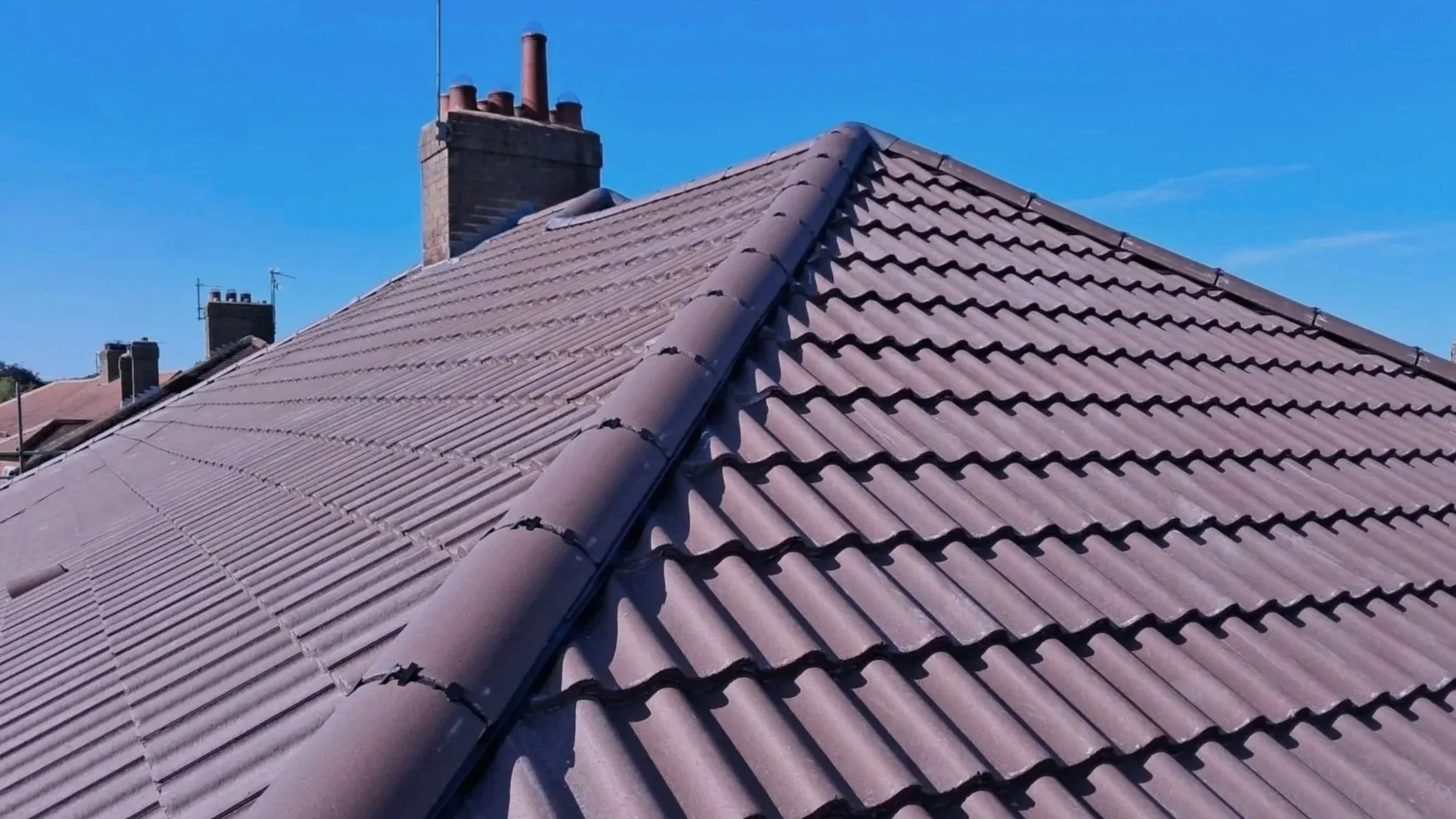 Close-up of a rooftop with brown Marley tiles and a central hip ridge, with a chimneys against a clear blue sky.