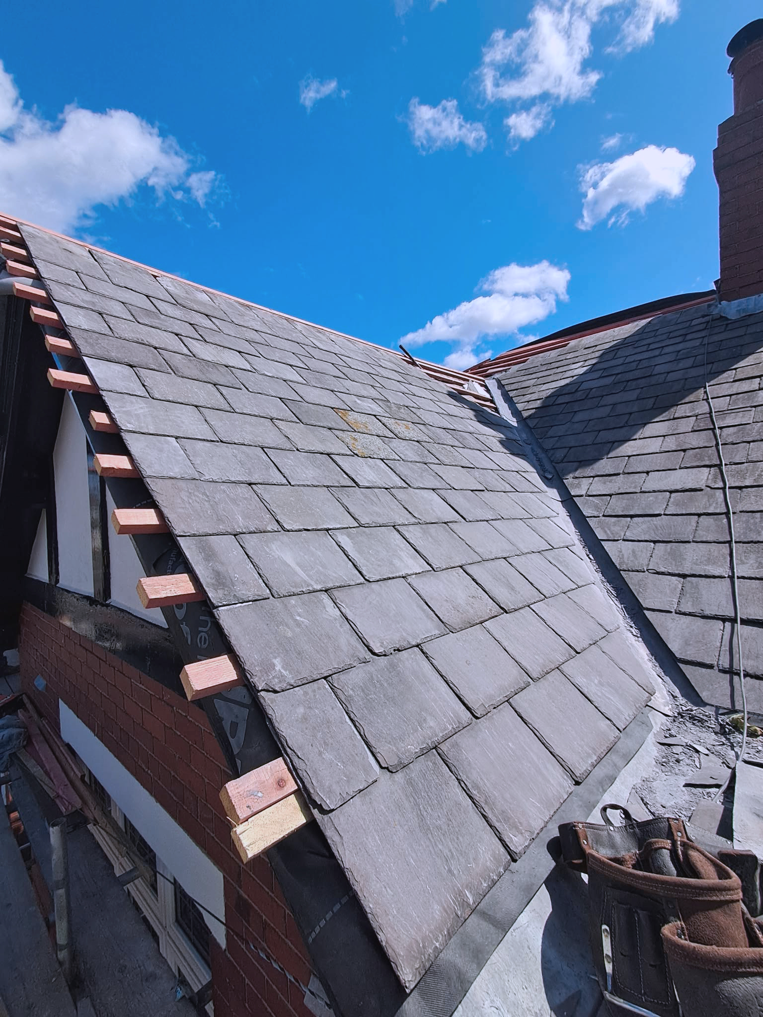 Construction site with roofing reclaimed slates on a sloped roof, partially unfinished, with a blue sky and clouds overhead, and a brick wall below.