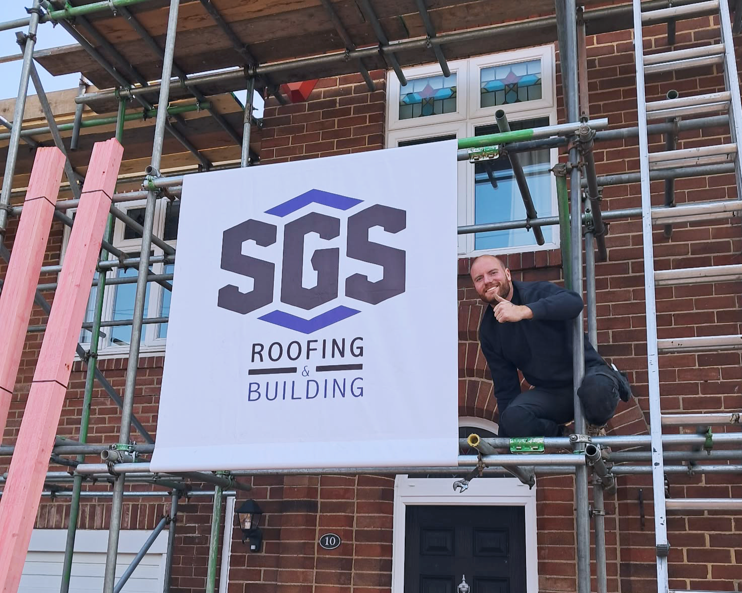 A man kneeling on scaffolding in front of a brick house, giving a thumbs up, next to a banner that reads 'SGS Roofing & Building'."