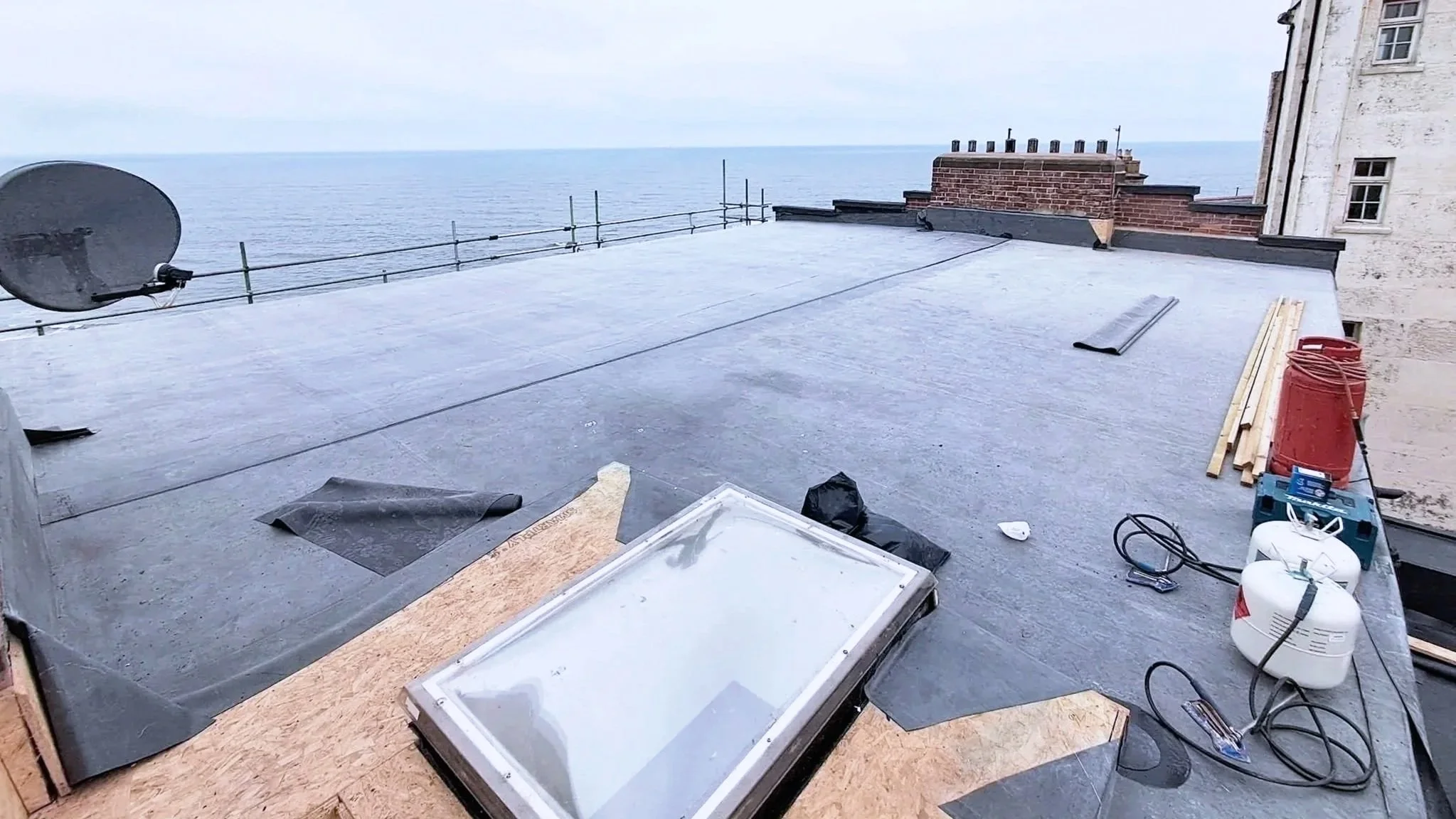 Rooftop under construction with tools, building materials, and a satellite dish, overlooking the ocean under an overcast sky.