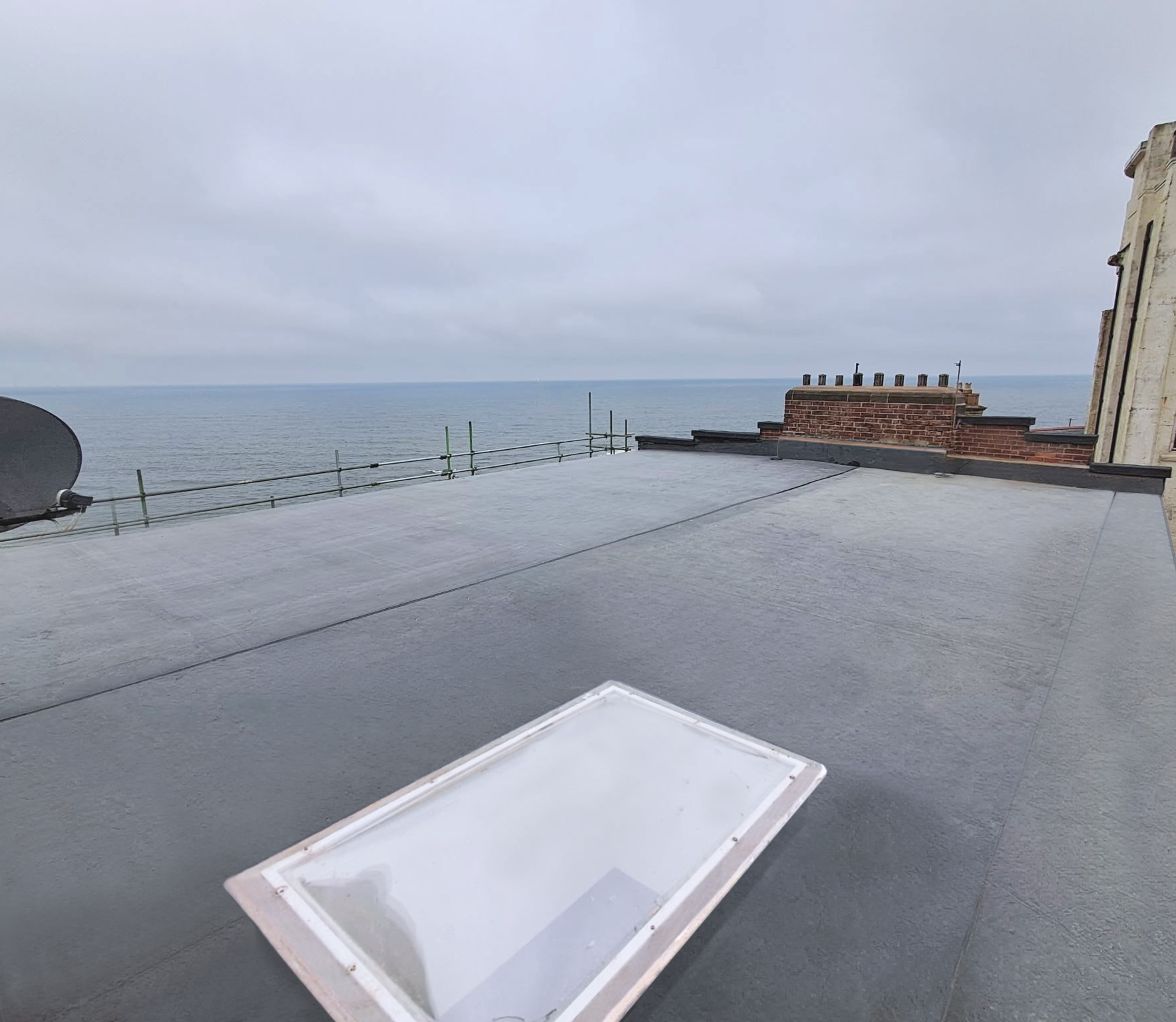 EPDM Rooftop view of the ocean on a cloudy day, with a chimney and a satellite dish on the roof, and a white tray or box in the foreground.