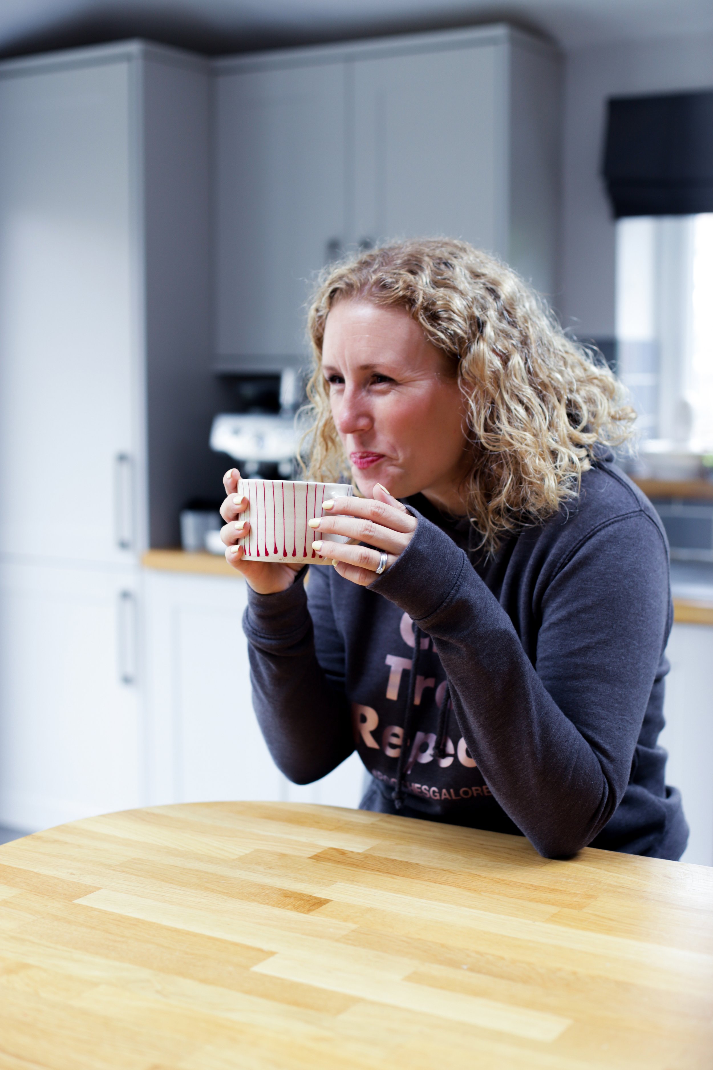 A woman with curly blonde hair wearing a dark hoodie is sitting at a wooden table, holding a white mug with red vertical stripes and enjoying a hot beverage in a kitchen setting.